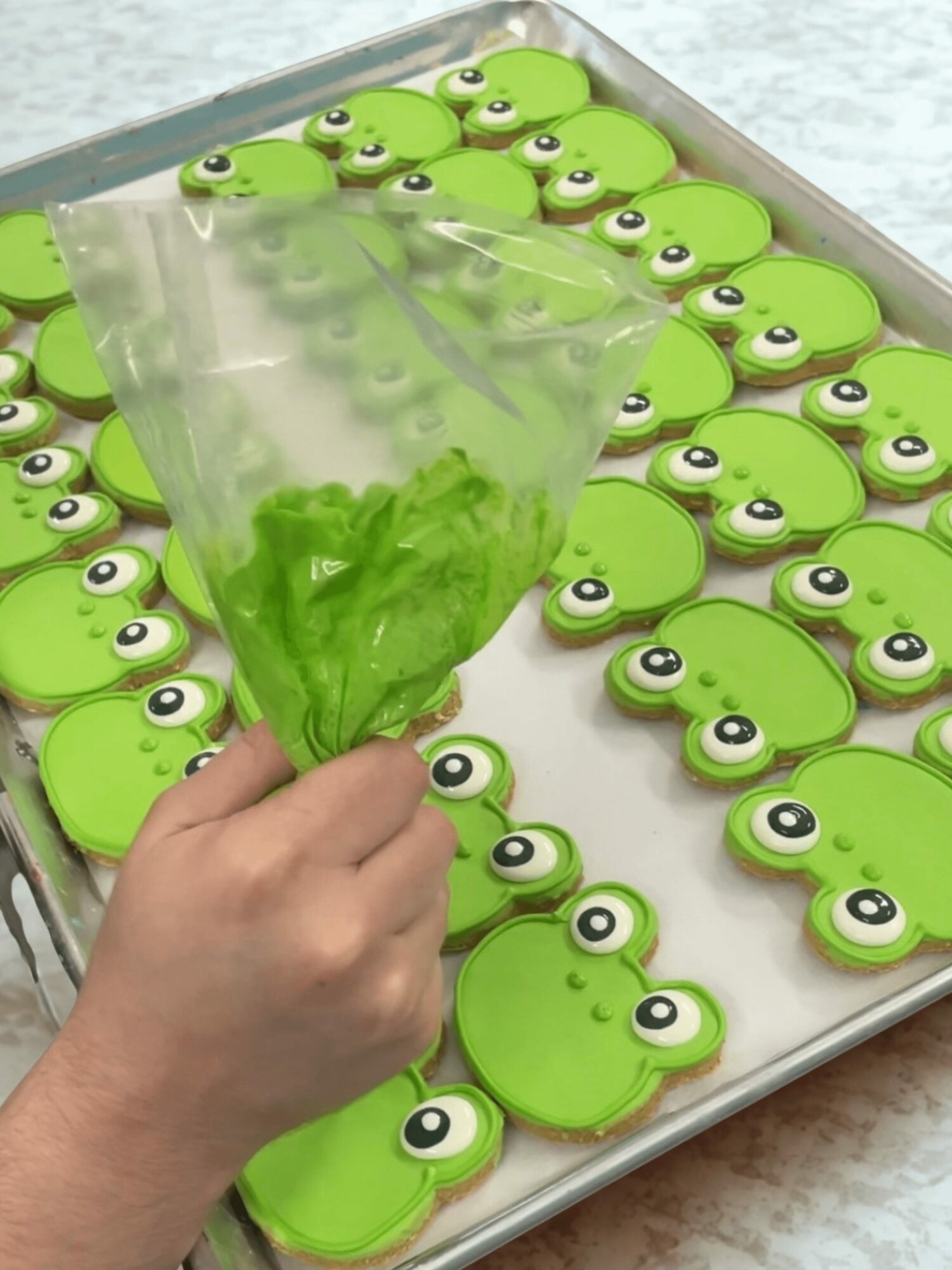 Hand holding a clear bag filled with green candies, surrounded by frog-shaped cookies with big eyes on a tray.