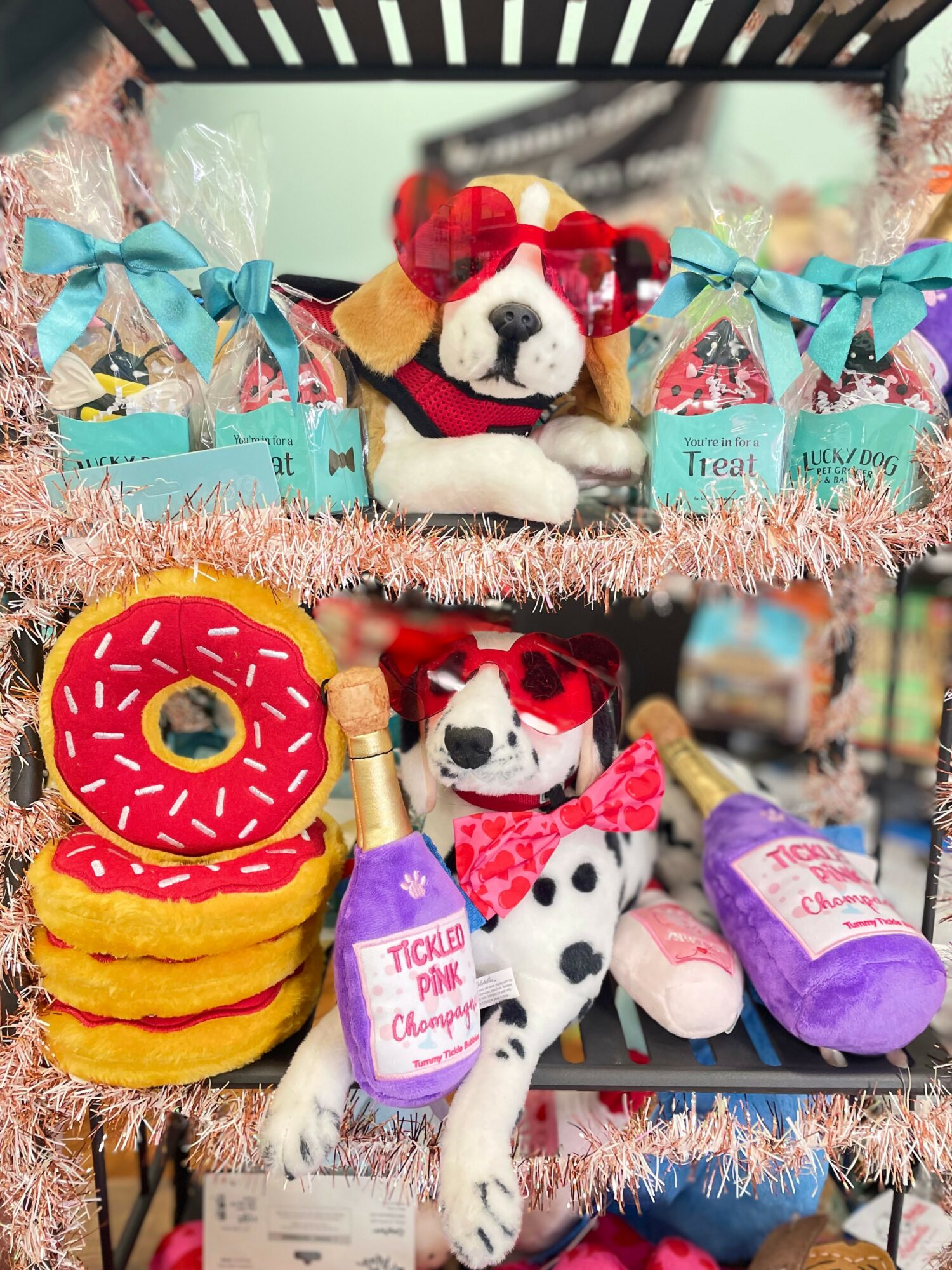 Decorative display with plush Dalmatian dogs wearing sunglasses and scarves, colorful toys, and a pink donut-shaped item, set on a pink tinsel background.