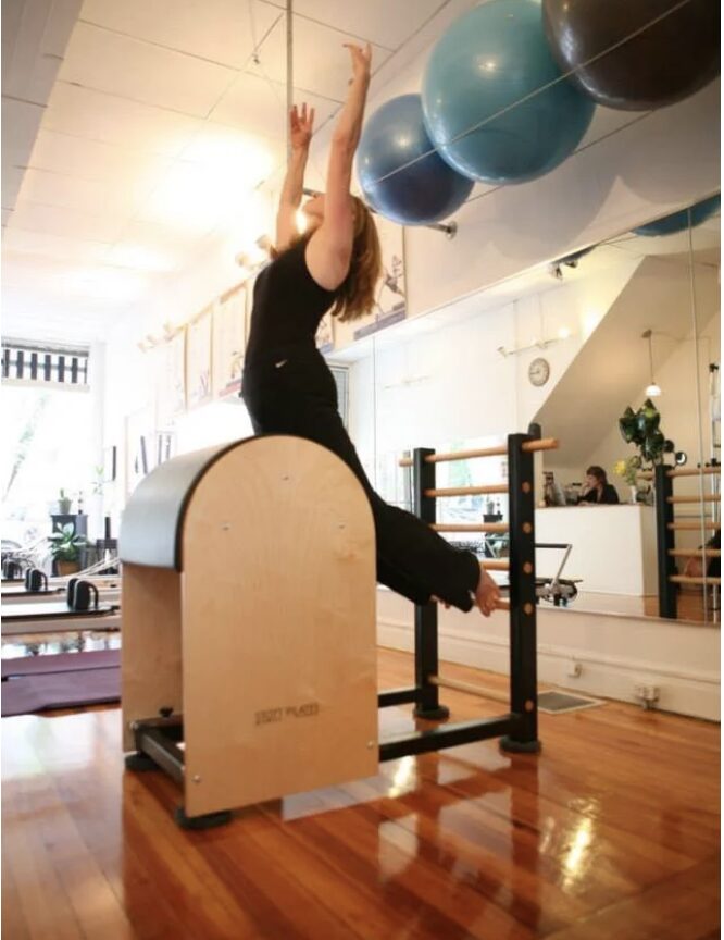 Person stretching with arms raised, sitting on a wooden exercise equipment in a bright room with exercise balls overhead.