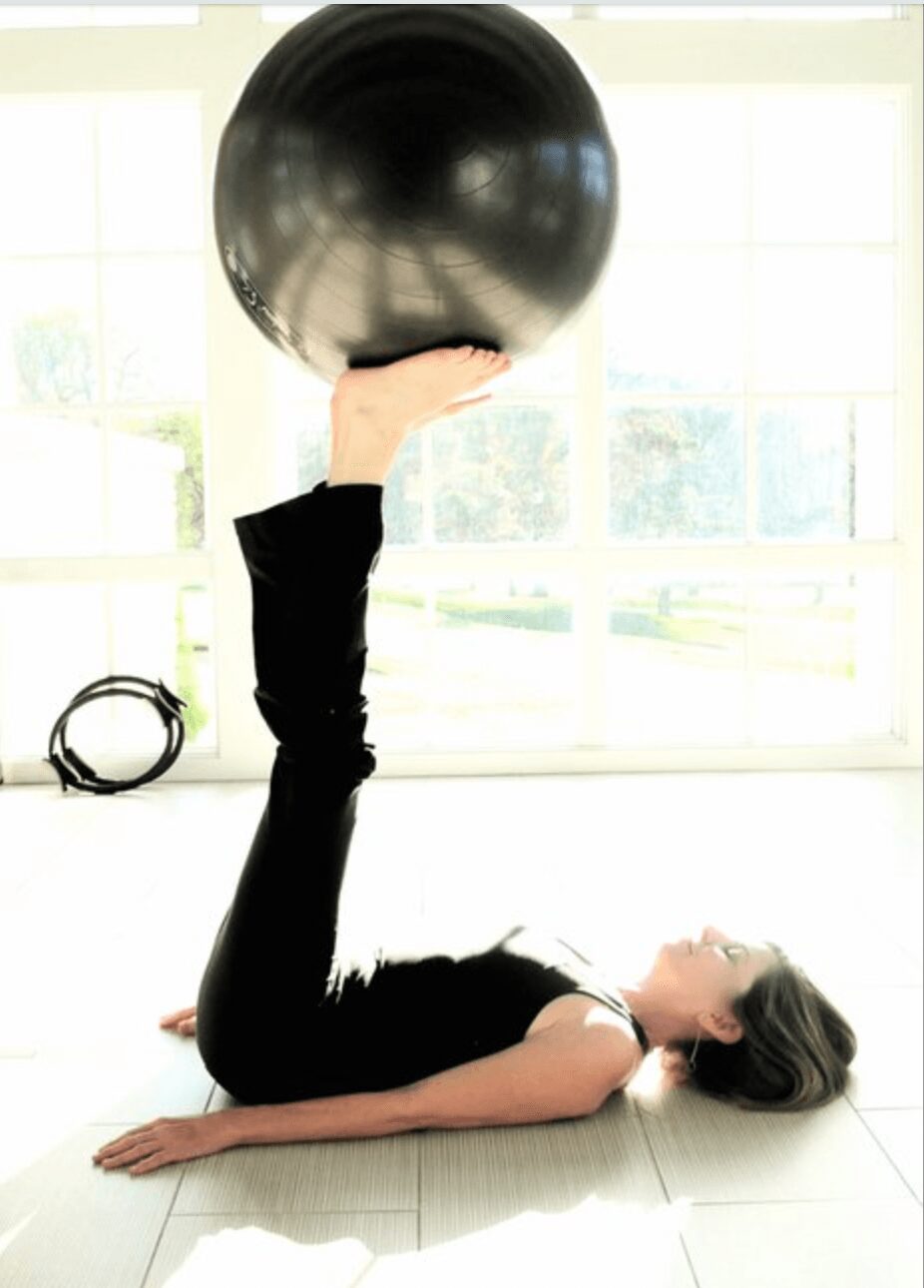 Woman lying on her back on the floor, holding a large black exercise ball above her with one hand, in a bright room.