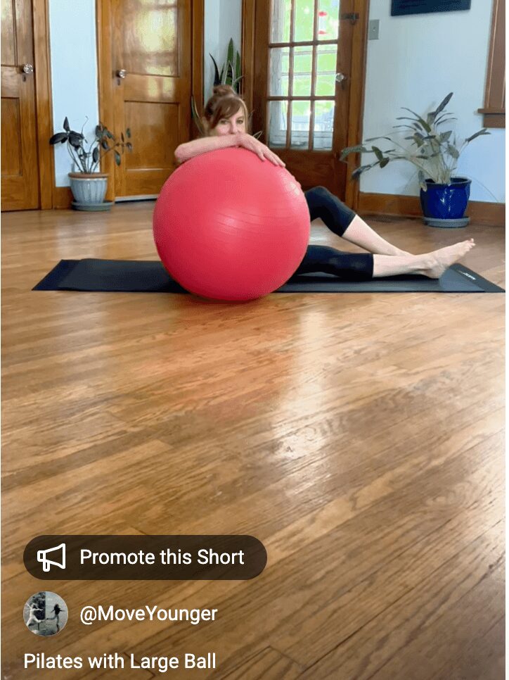 Child lying on a yoga mat, hugging a large red exercise ball in a room with wooden floors and potted plants.