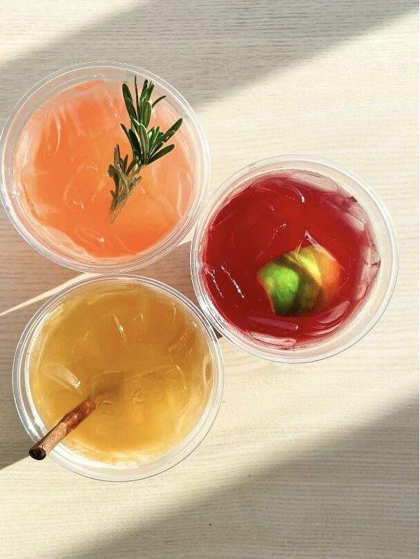 Three glasses of colorful beverages with ice, garnished with herbs and fruit slices, on a light surface.