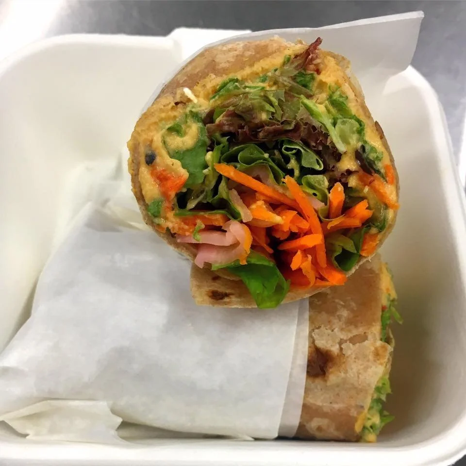 Half of a vegetable wrap with lettuce, carrots, and other vegetables inside a pita, partially wrapped in paper, in a white container.