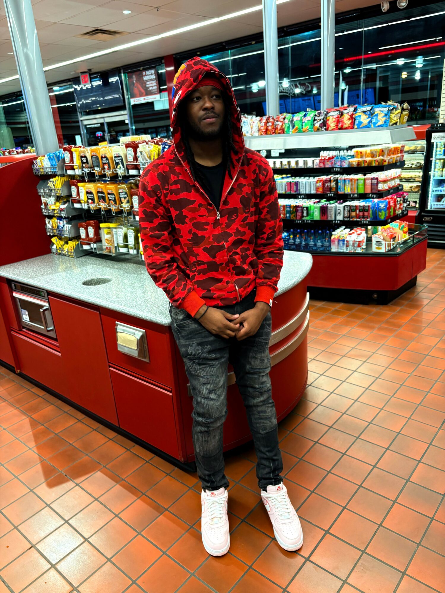 Person in red hoodie and gray pants standing inside a store with shelves of snacks behind them.