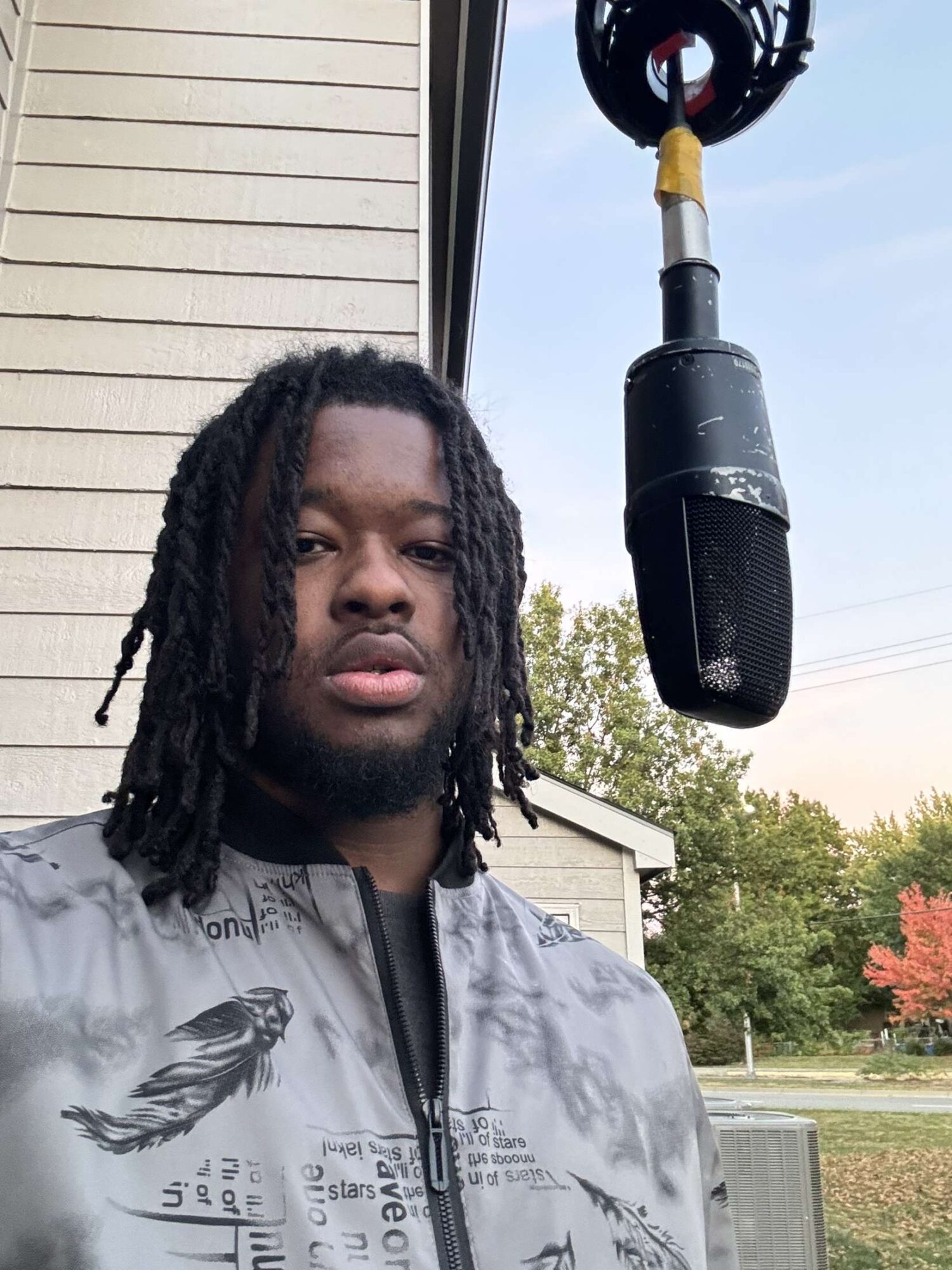 Young man with dreadlocks standing outdoors near a building, microphone hanging from above, trees in background.