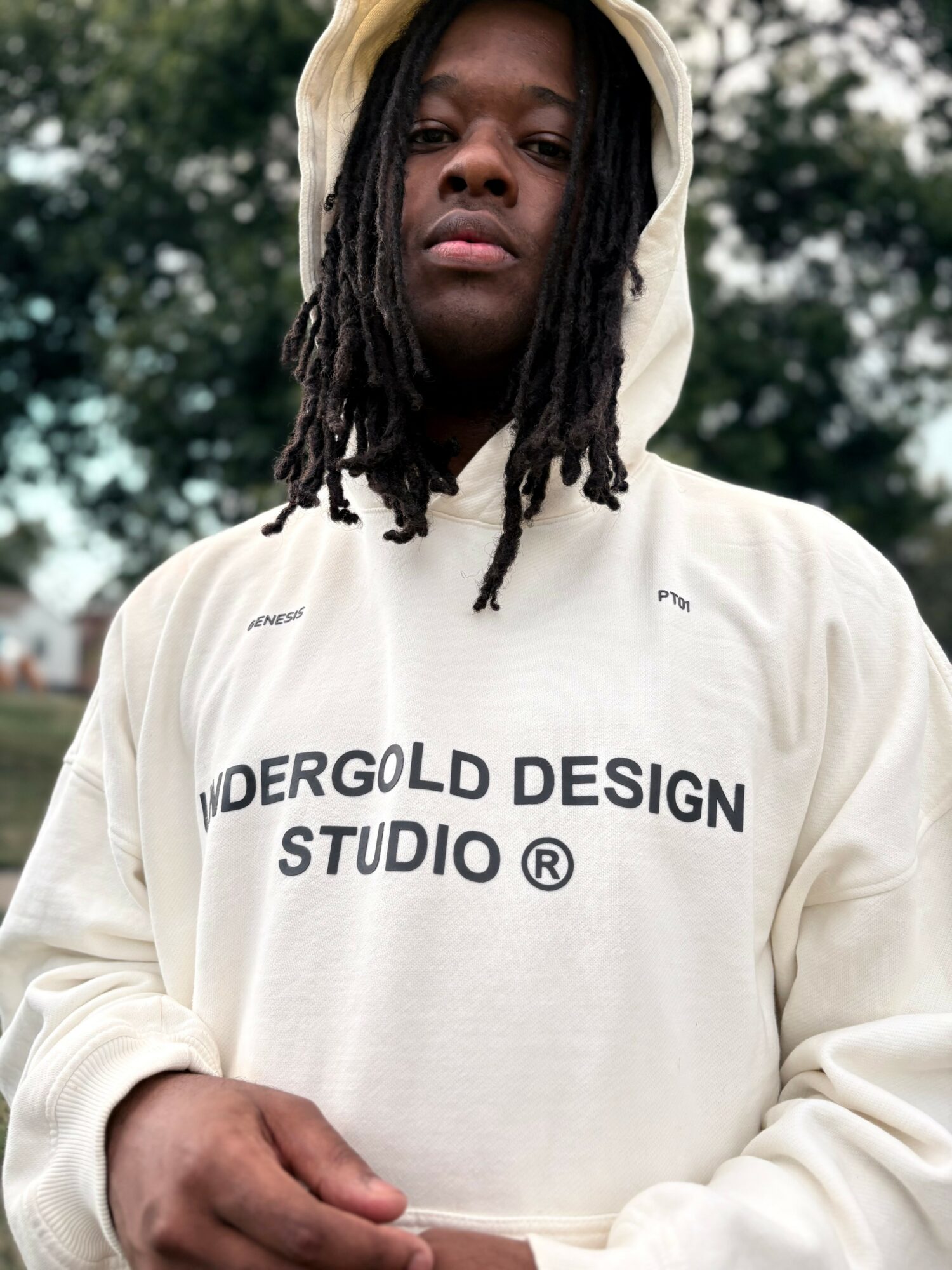 Young person with dreadlocks wearing a white hoodie with text, outdoors with trees in background.