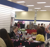 People sitting at tables in a large indoor space, some using laptops, with booths and displays in the background.