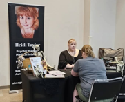 Two women sit at a table with promotional materials and a banner displaying a woman's face and text, in an indoor setting.
