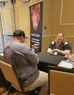 Two people sitting at a table in a conference room, one smiling and the other with back to camera, with a banner behind them.