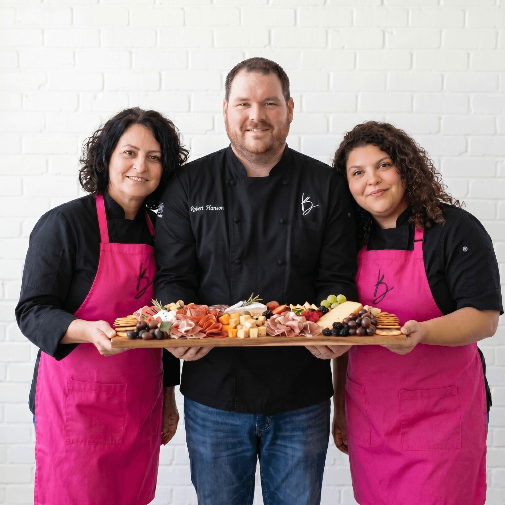 Three people, two women and one man, stand together holding a large tray of assorted food, smiling, against a white brick wall.