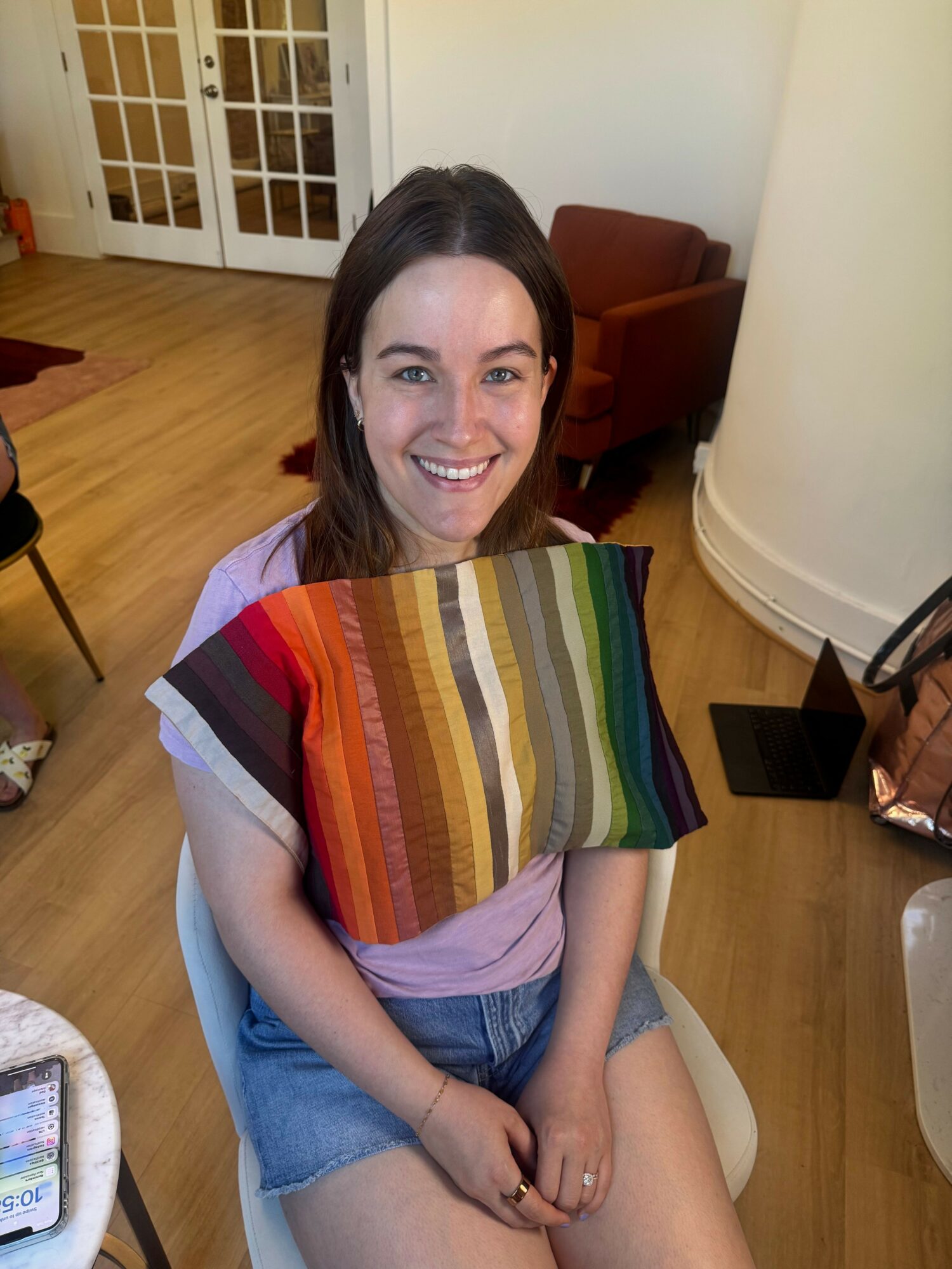 Young woman sitting on a chair, smiling, wearing a colorful striped top and denim shorts, in a room with wooden floor.