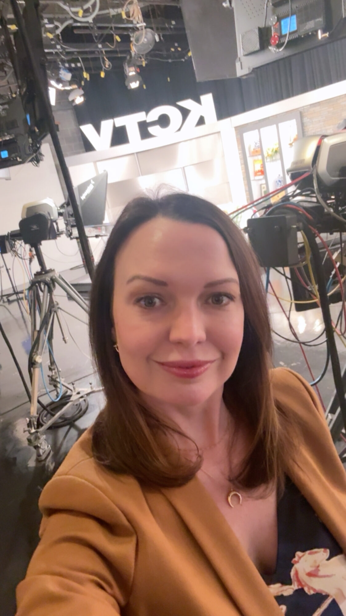 Woman with brown hair and a tan blazer taking a selfie in a studio with cameras and equipment.
