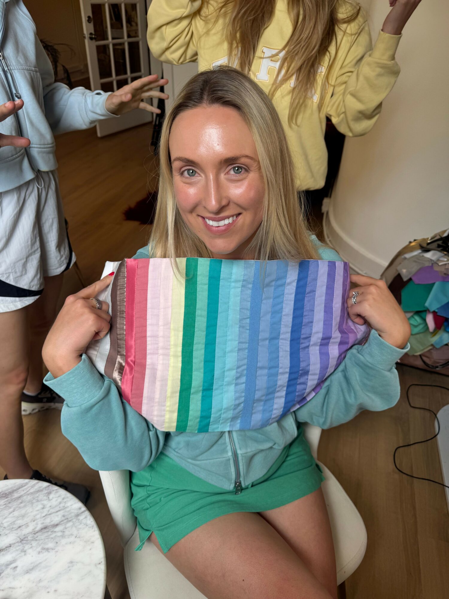 Woman sitting on a chair holding a colorful striped object, smiling, with a person in the background reaching out.