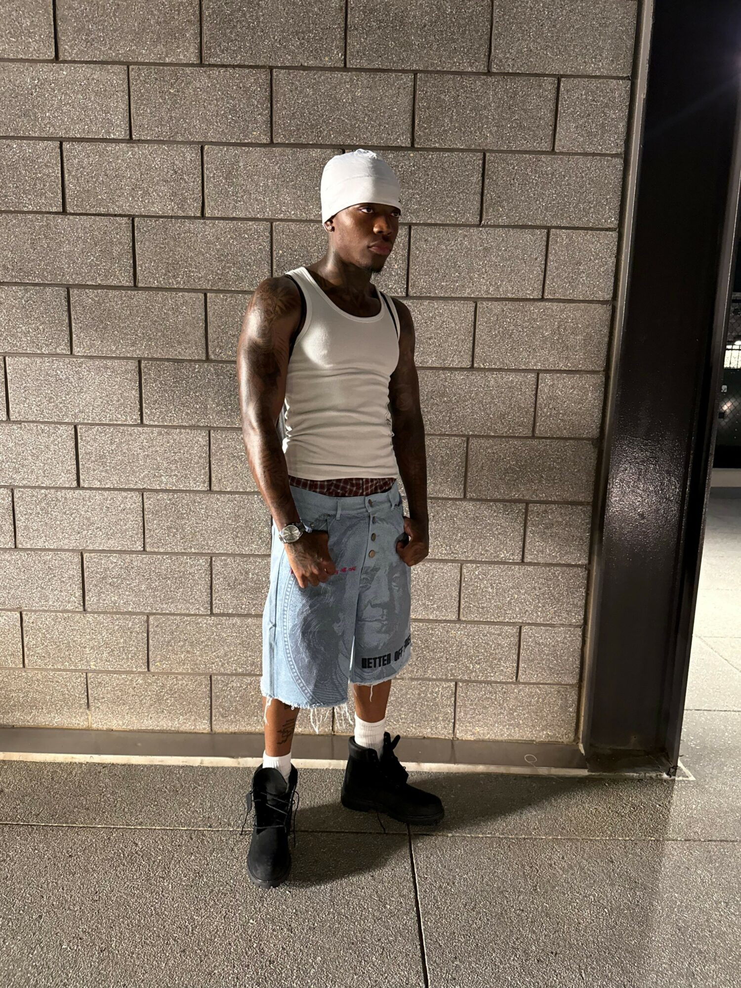 Person standing against a brick wall, wearing a white tank top, denim shorts, and black shoes, with a white cap.
