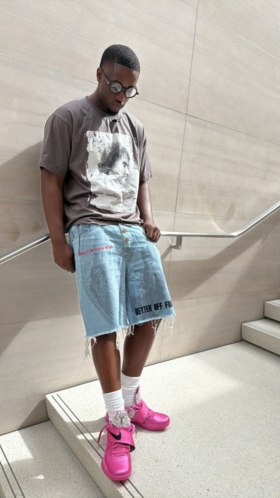 Young person standing on stairs, wearing glasses, a graphic t-shirt, denim shorts, white socks, and pink sneakers.