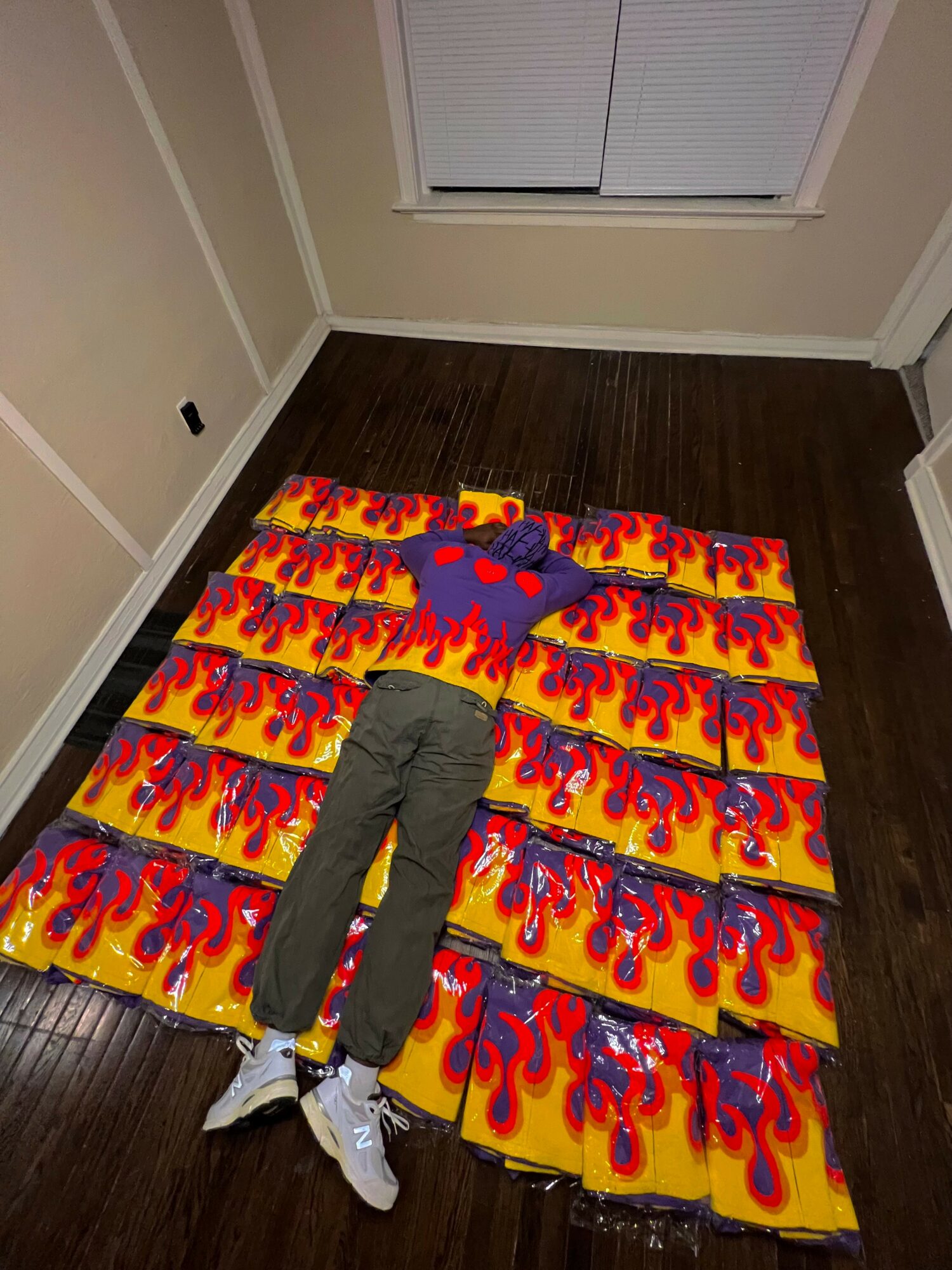 Person lying on colorful skateboard decks with flame design, arranged on dark wooden floor in room with window and beige walls.