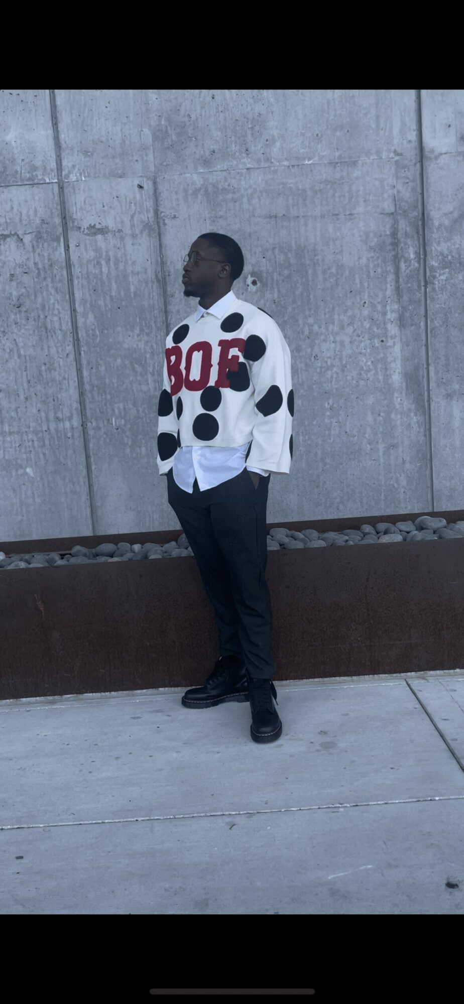 Person standing sideways against a concrete wall, wearing a polka dot jacket with red and black lettering, dark pants, and shoes.