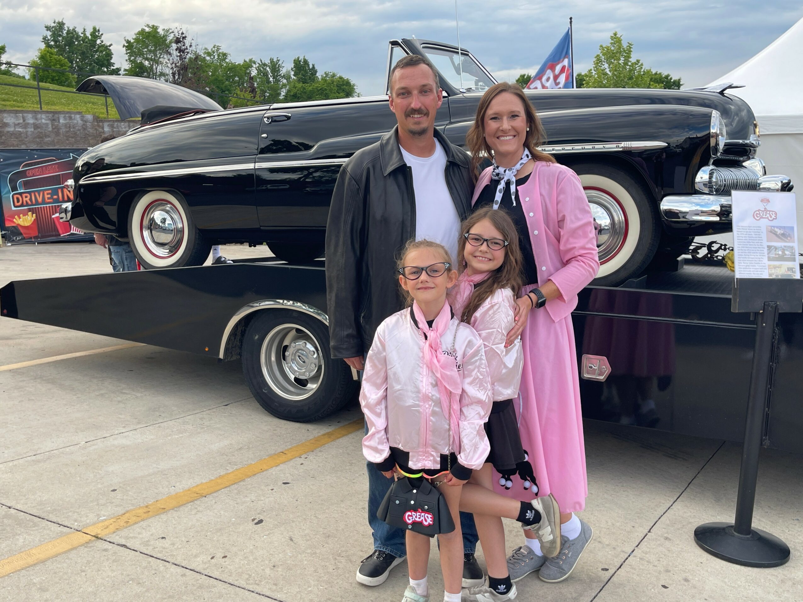 Family of four standing outdoors near a vintage black car on a trailer, with trees and a cloudy sky in the background.