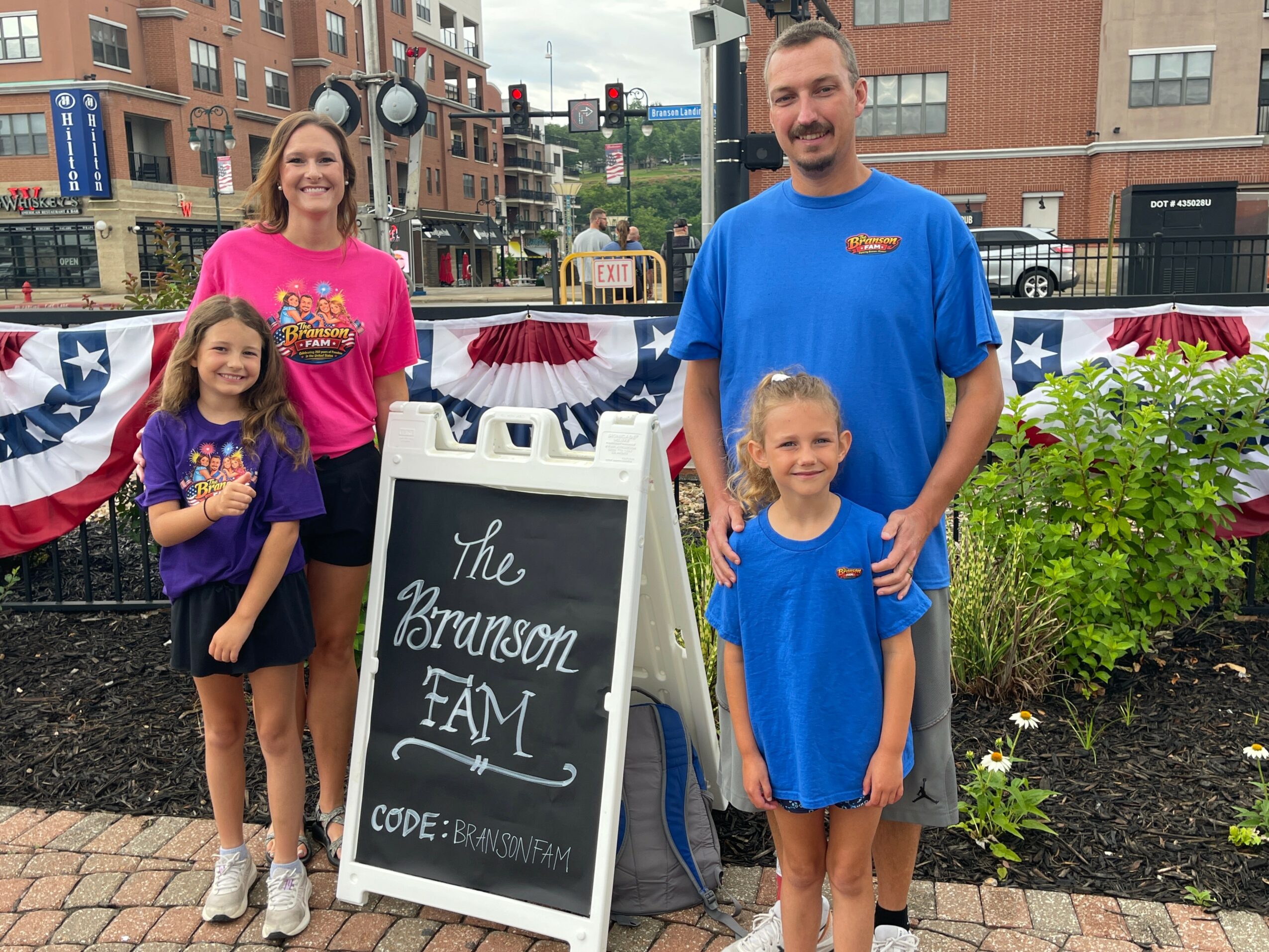 Family of four standing outdoors with a chalkboard sign, decorated with red, white, and blue bunting, in a city park.