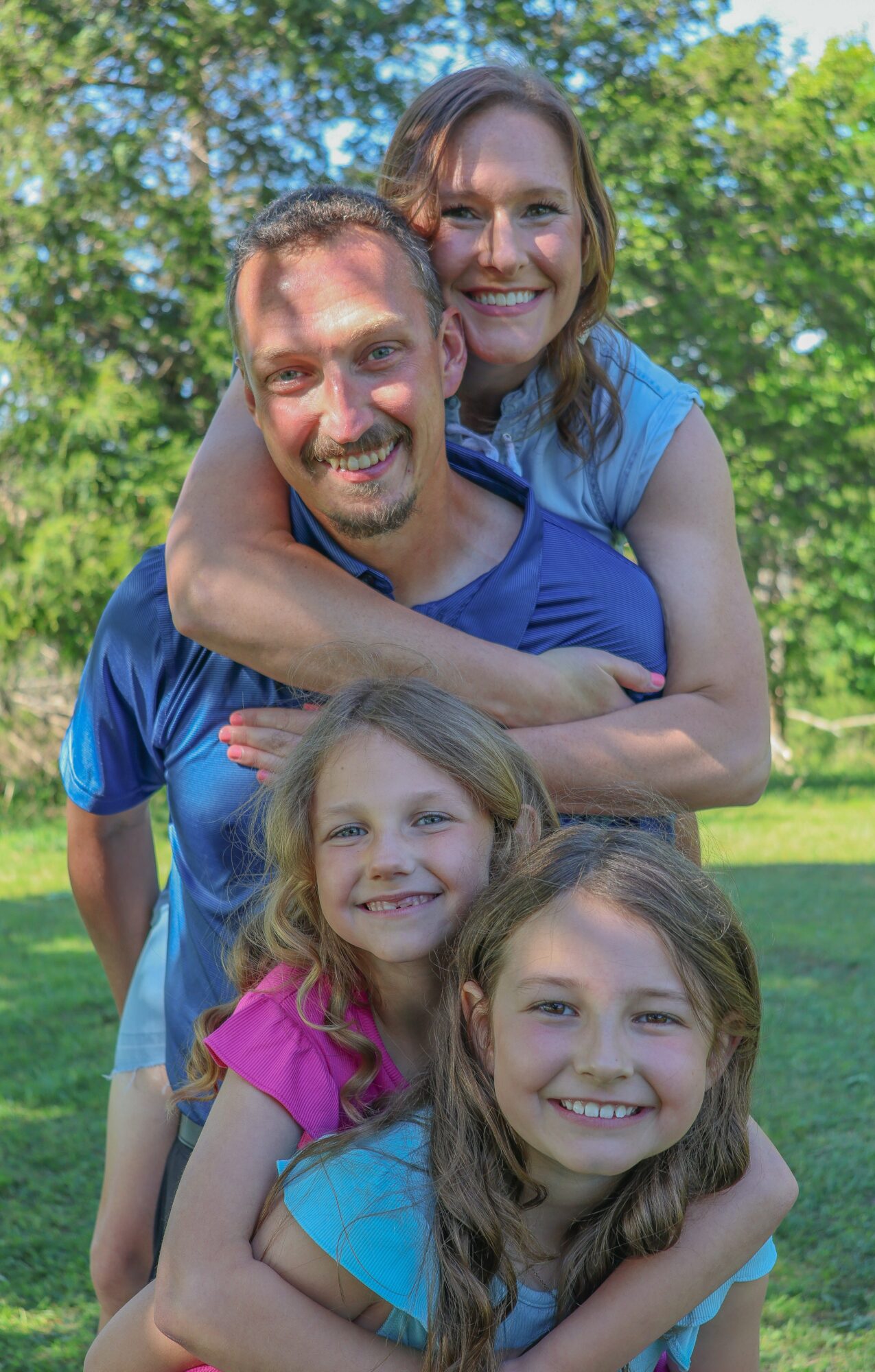 Four people outdoors, smiling, with trees and blue sky in background.