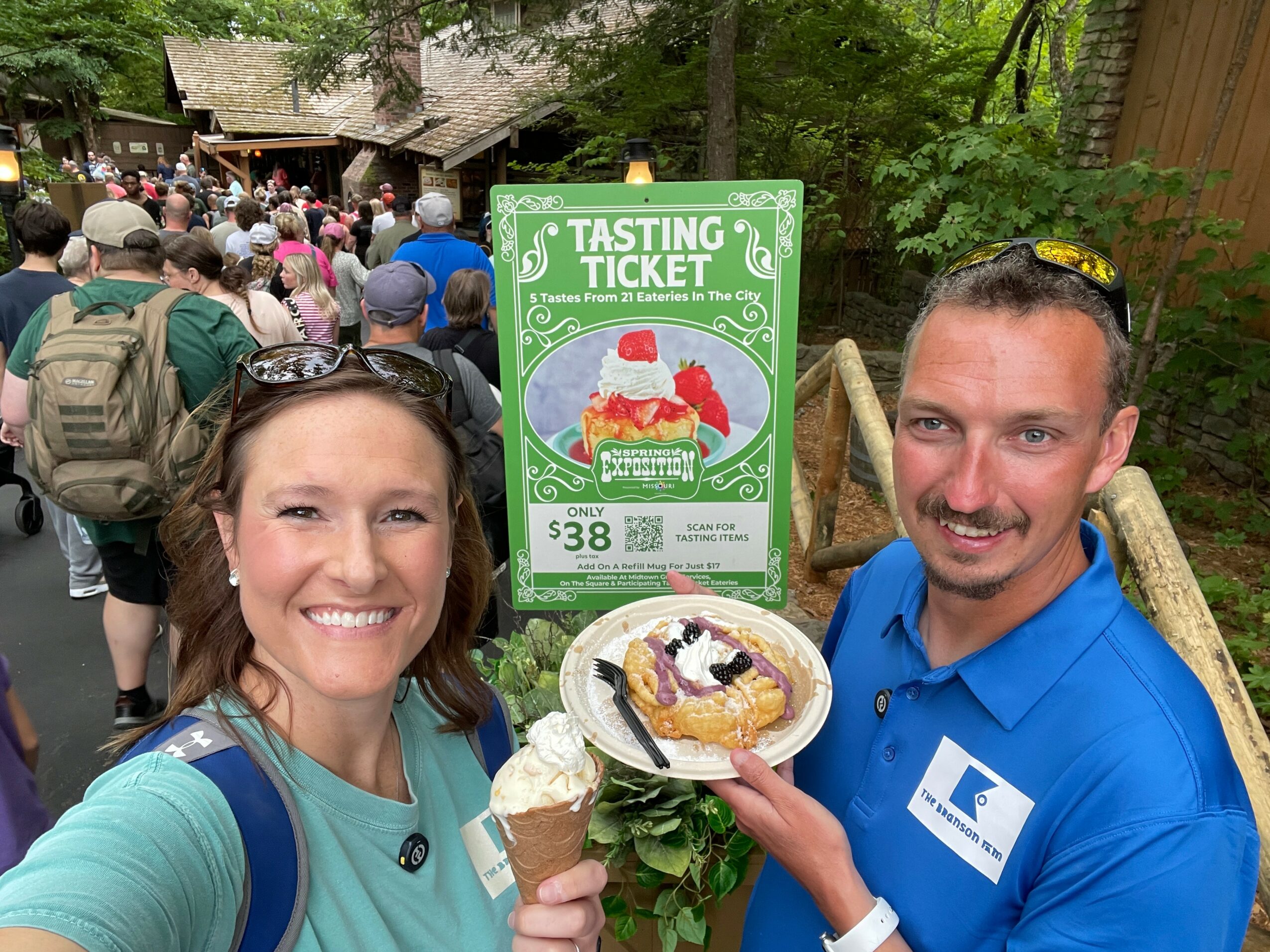 Two smiling people holding a plate with a dessert and an ice cream cone at an outdoor event, with a crowd and a green sign in the background.