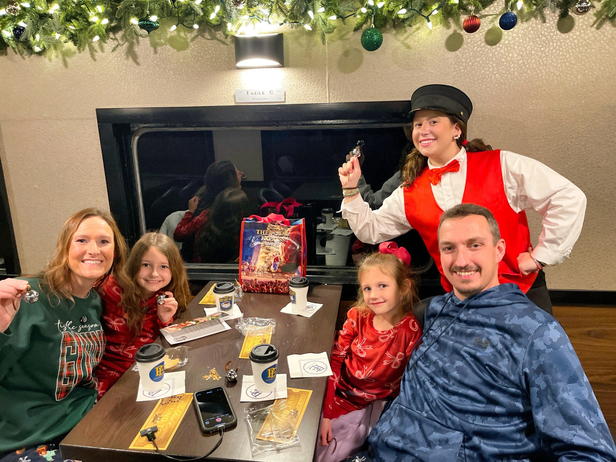 Group of five people, including children and an adult, sitting at a table with holiday decorations and a performer in a red vest and black hat, smiling.