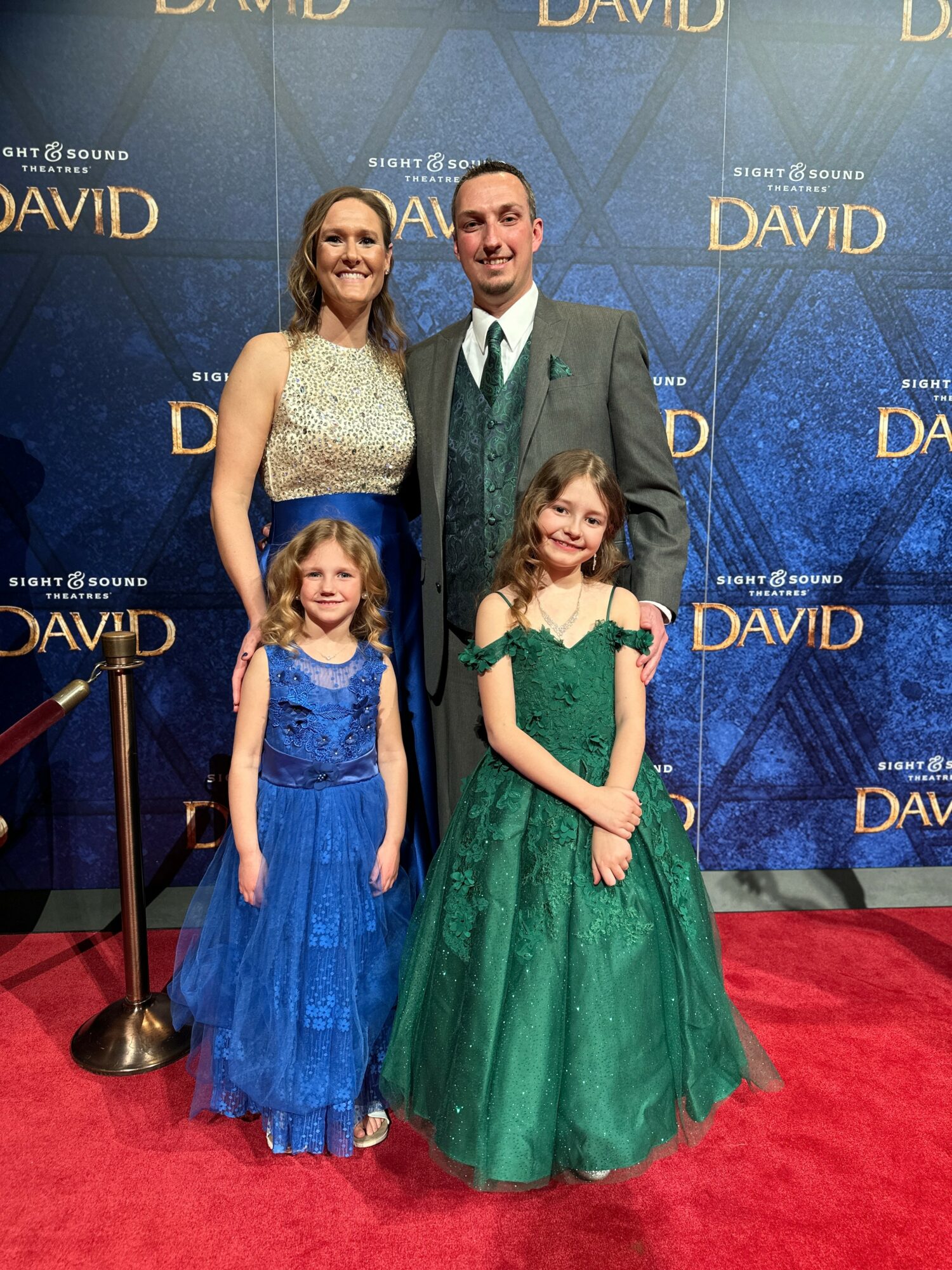Four people, two adults and two children, pose on a red carpet in front of a blue backdrop with gold text, smiling.