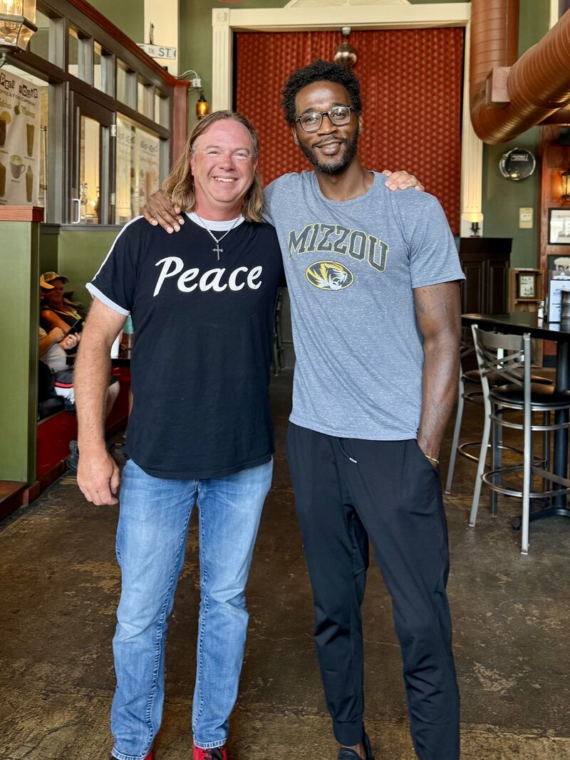 Two smiling men stand together in a restaurant, one wearing a black shirt with 'Peace' and the other in a Missouri T-shirt, with arms around each other.