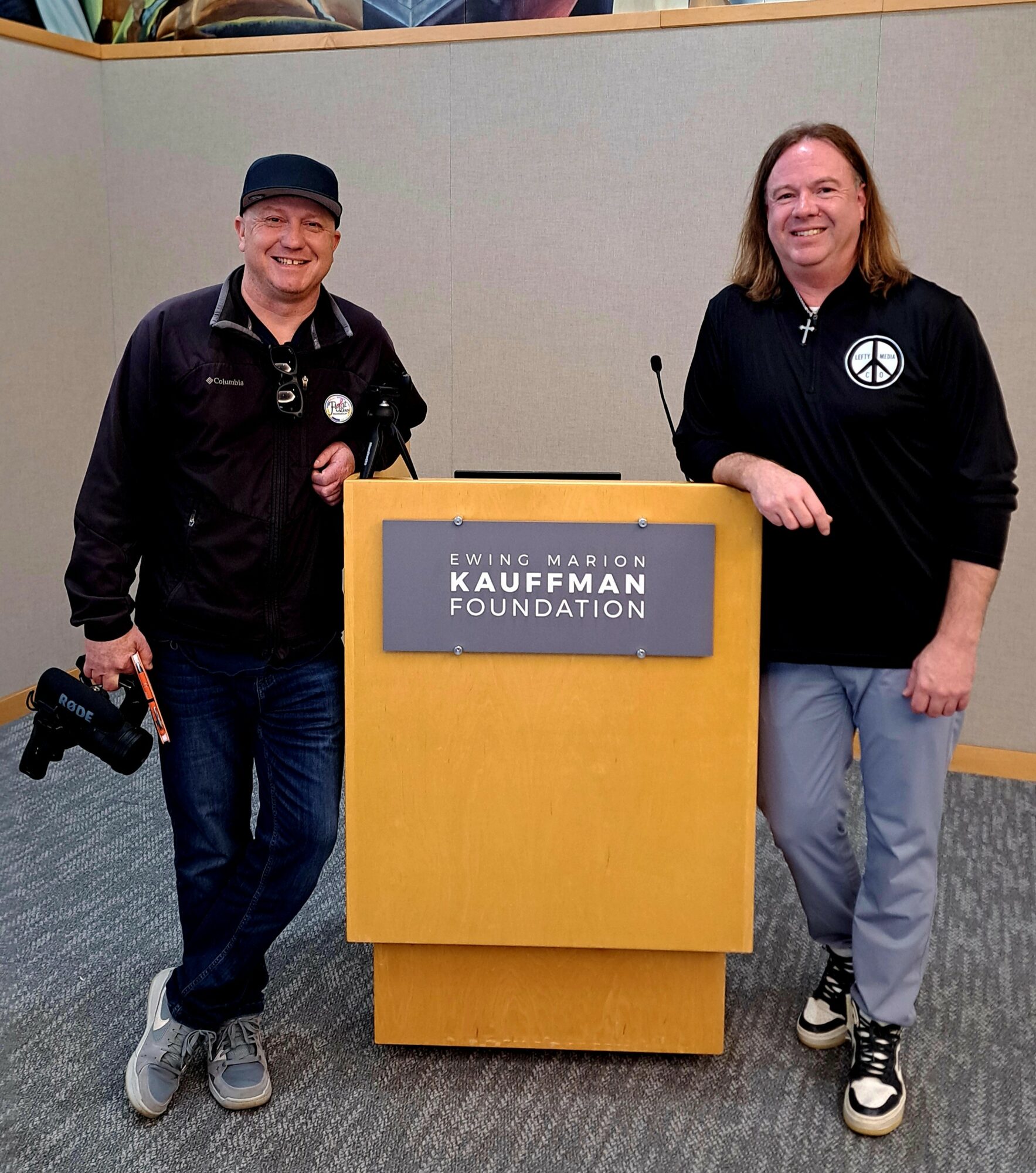Two people standing beside a podium with a sign reading 'Kauffman Foundation' in a room with gray walls.