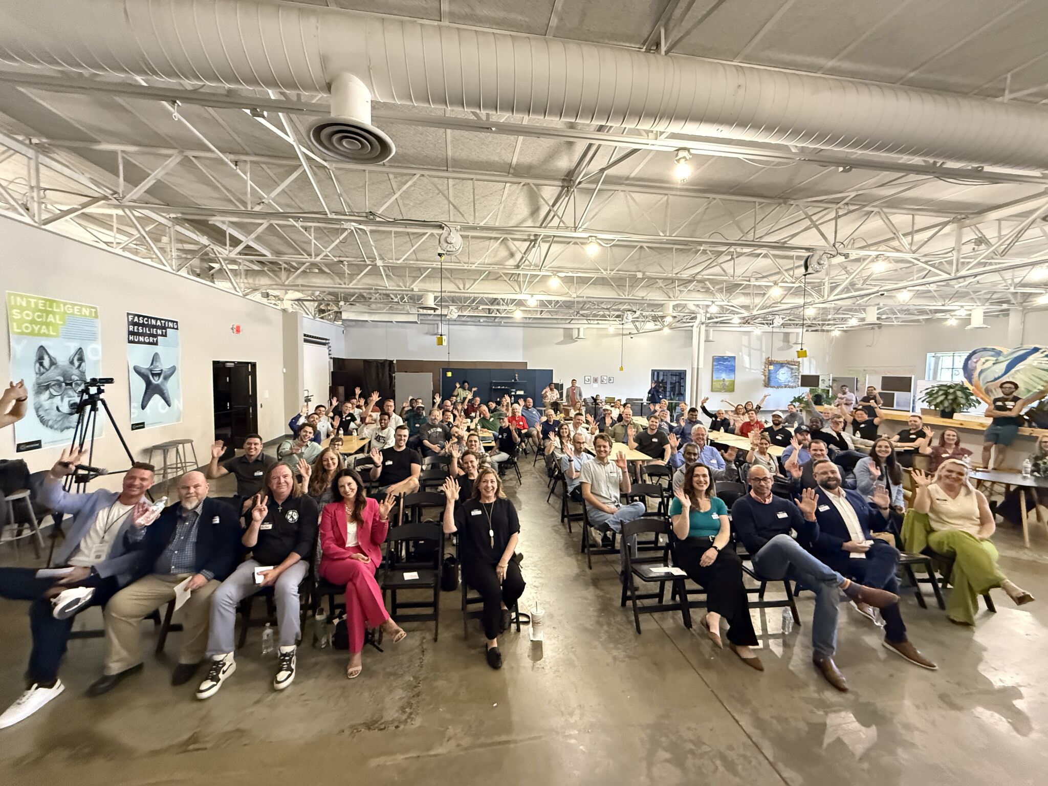 Large group of people seated in rows inside a spacious industrial-style hall with high ceilings and exposed ductwork.