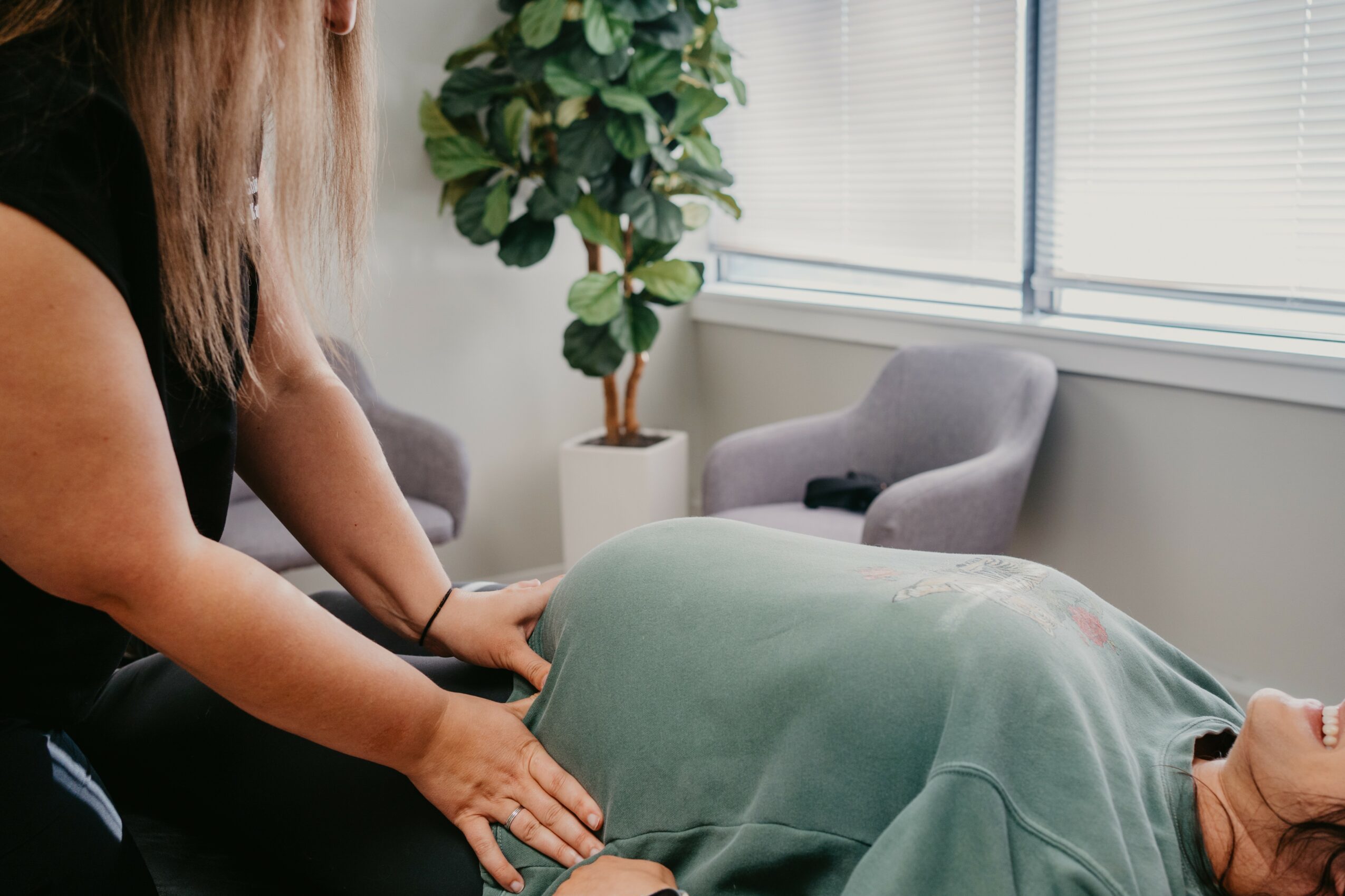 Person adjusting a green blanket on a bed in a bright room with chairs and a plant.