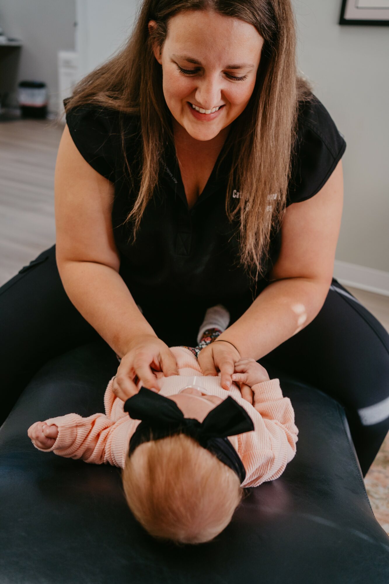 Woman smiling while holding a baby lying on a table, baby wearing a black bow headband, in a room with a gray wall.