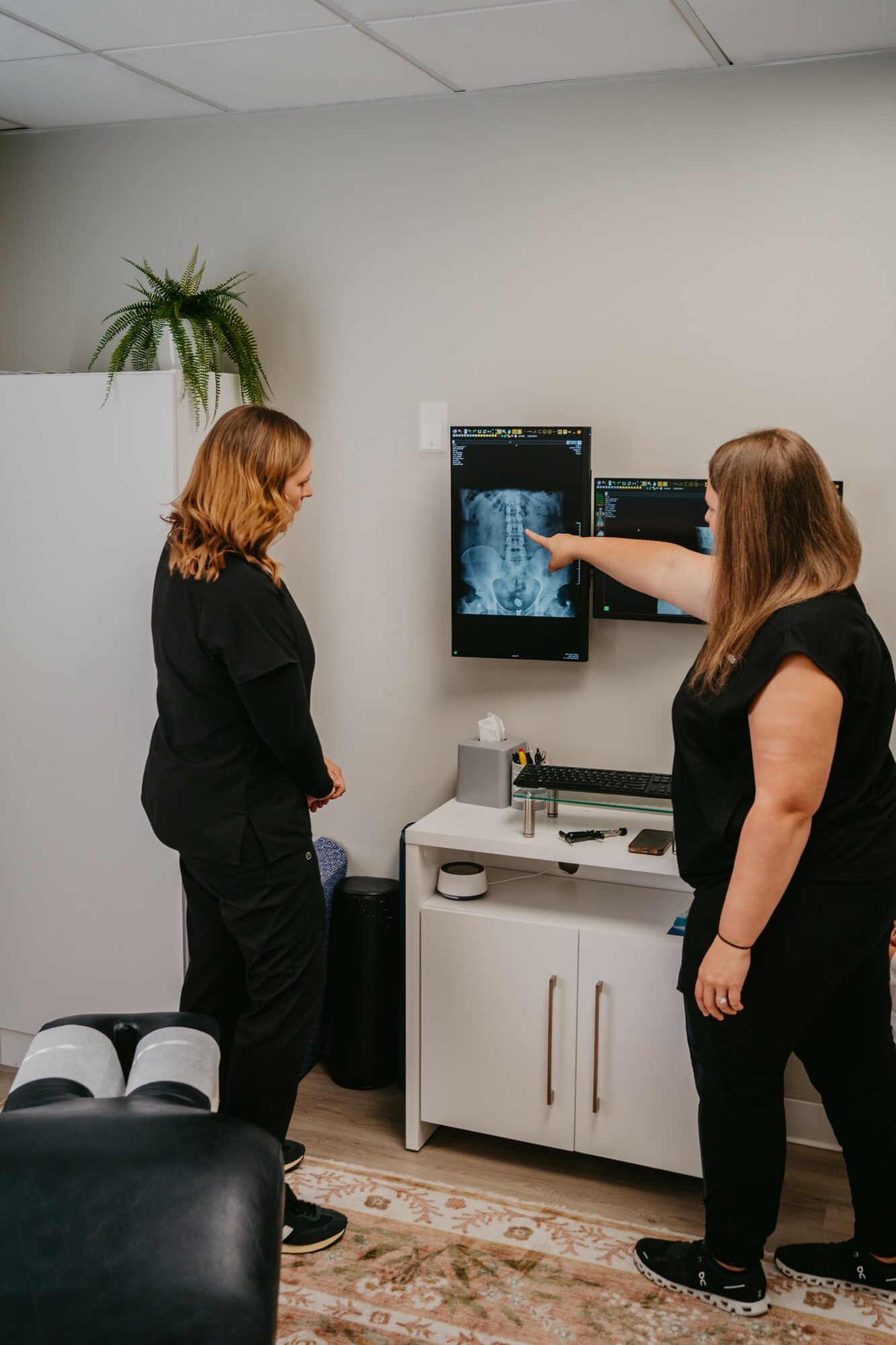Two women in black clothing discuss an X-ray image on a wall-mounted screen in a medical office, with a plant and white cabinet nearby.