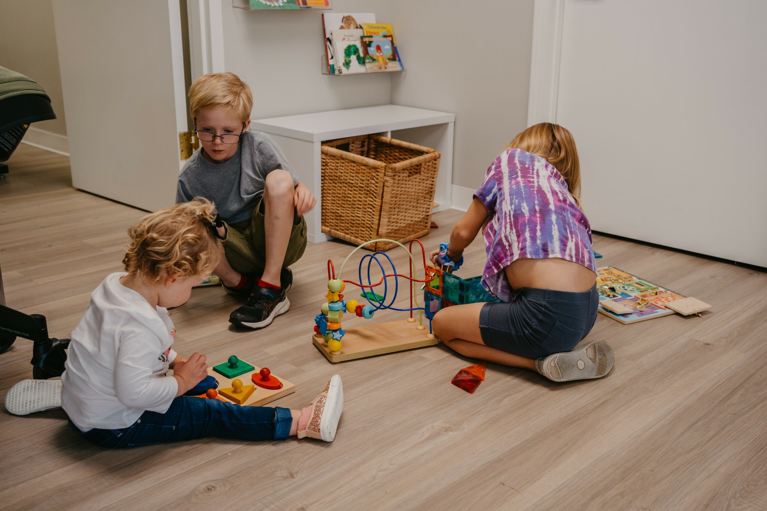 Two children playing with toys on a wooden floor in a room with white walls and a basket, one girl and one boy.