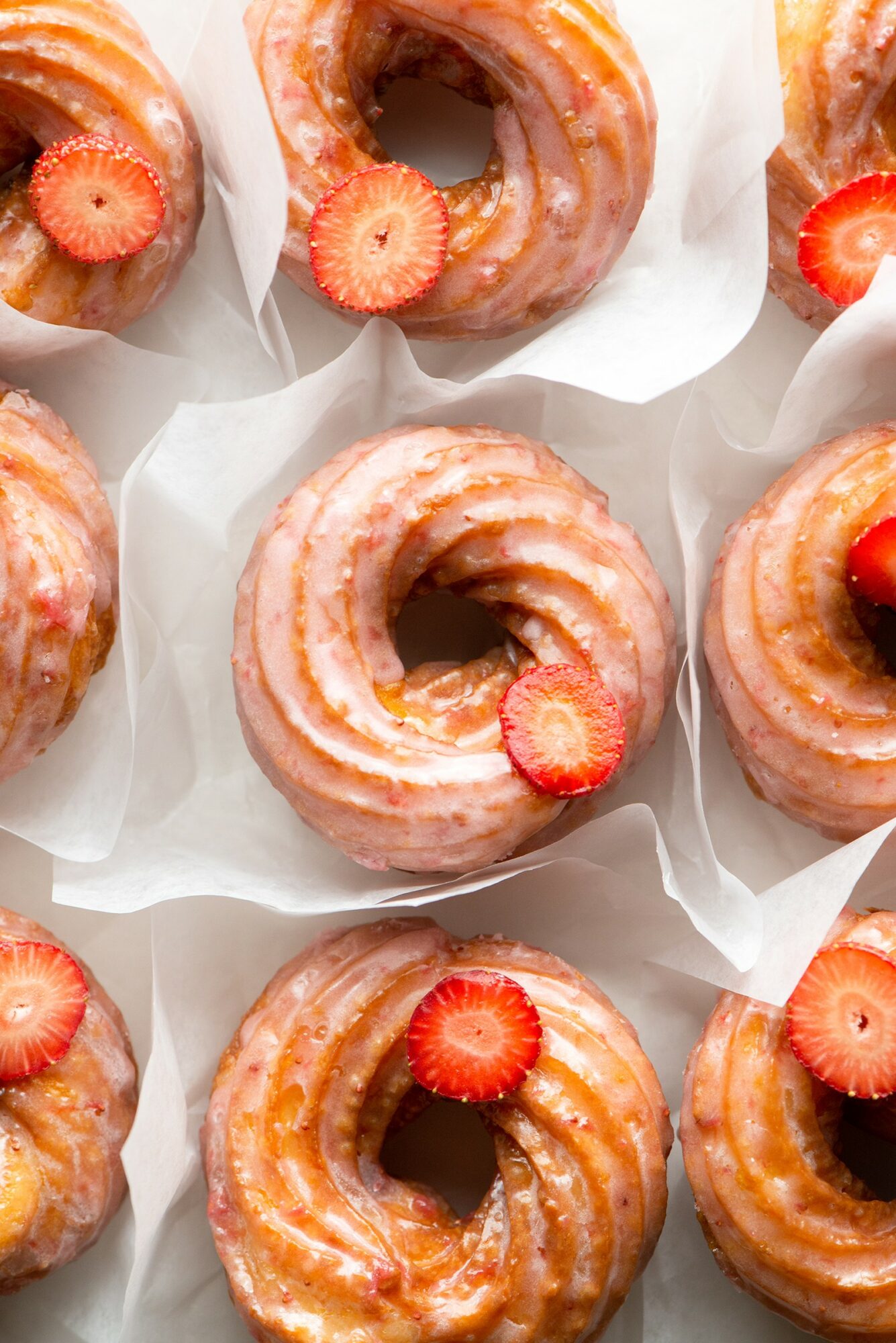 Six glazed donuts with strawberry slices on top, arranged on white paper wrappers, viewed from above.