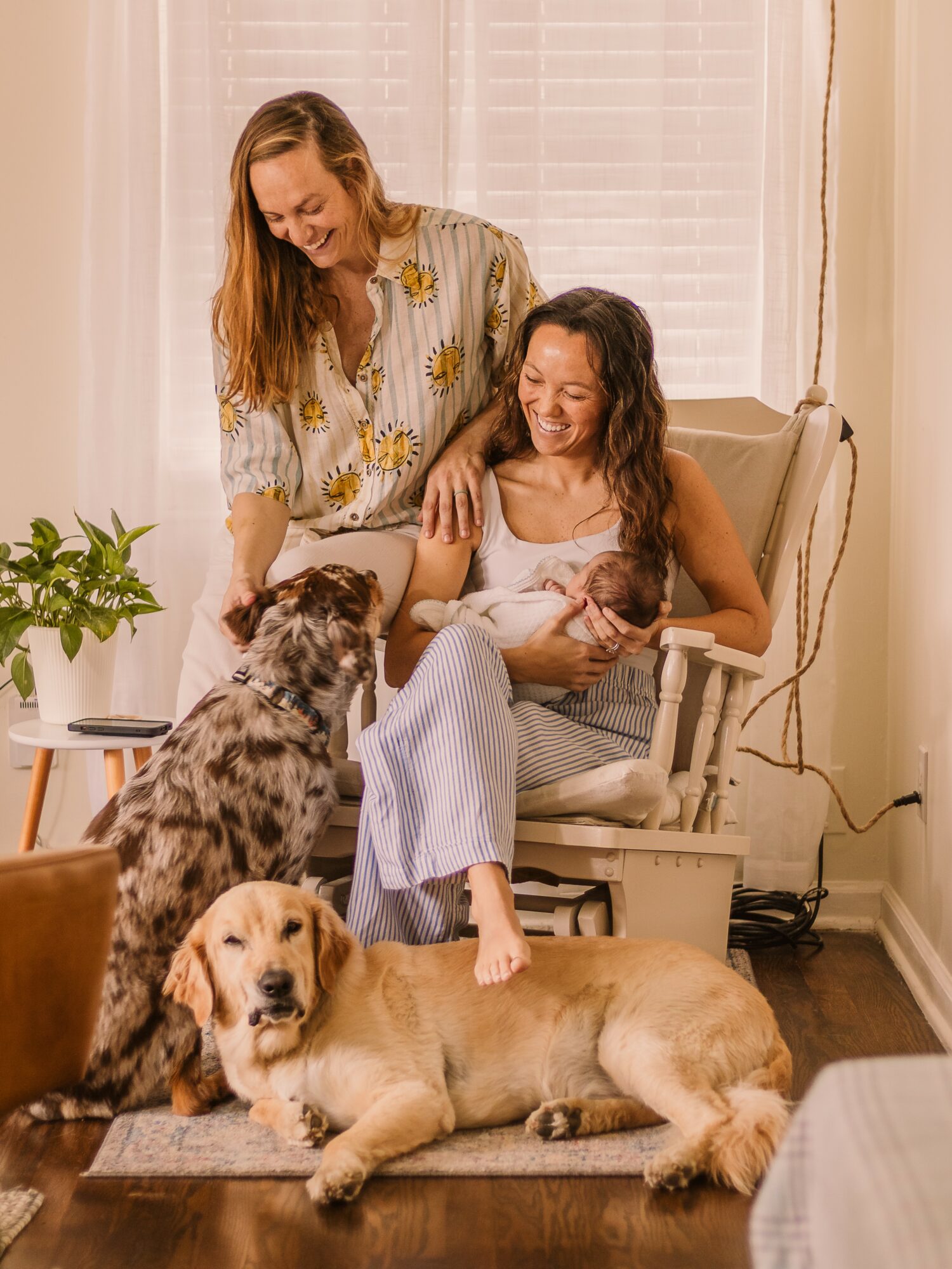 Two women and three dogs in a cozy room, one woman sitting in a chair, the other standing, all smiling.