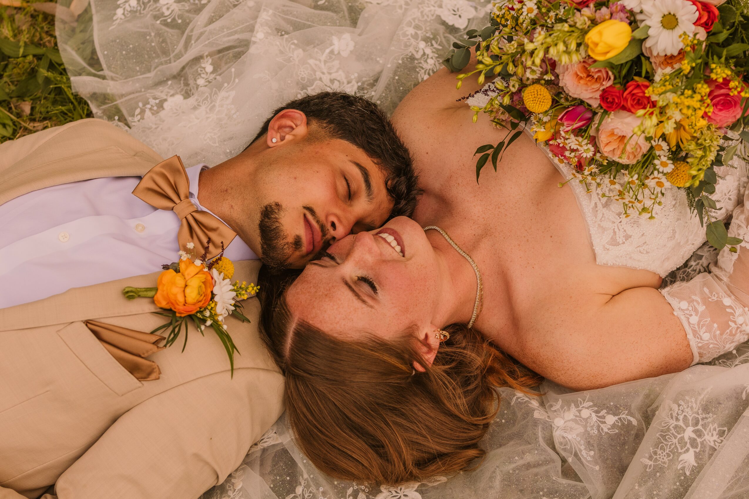 A couple lies on the ground, smiling and close together, with flowers on their clothing and a bouquet nearby.