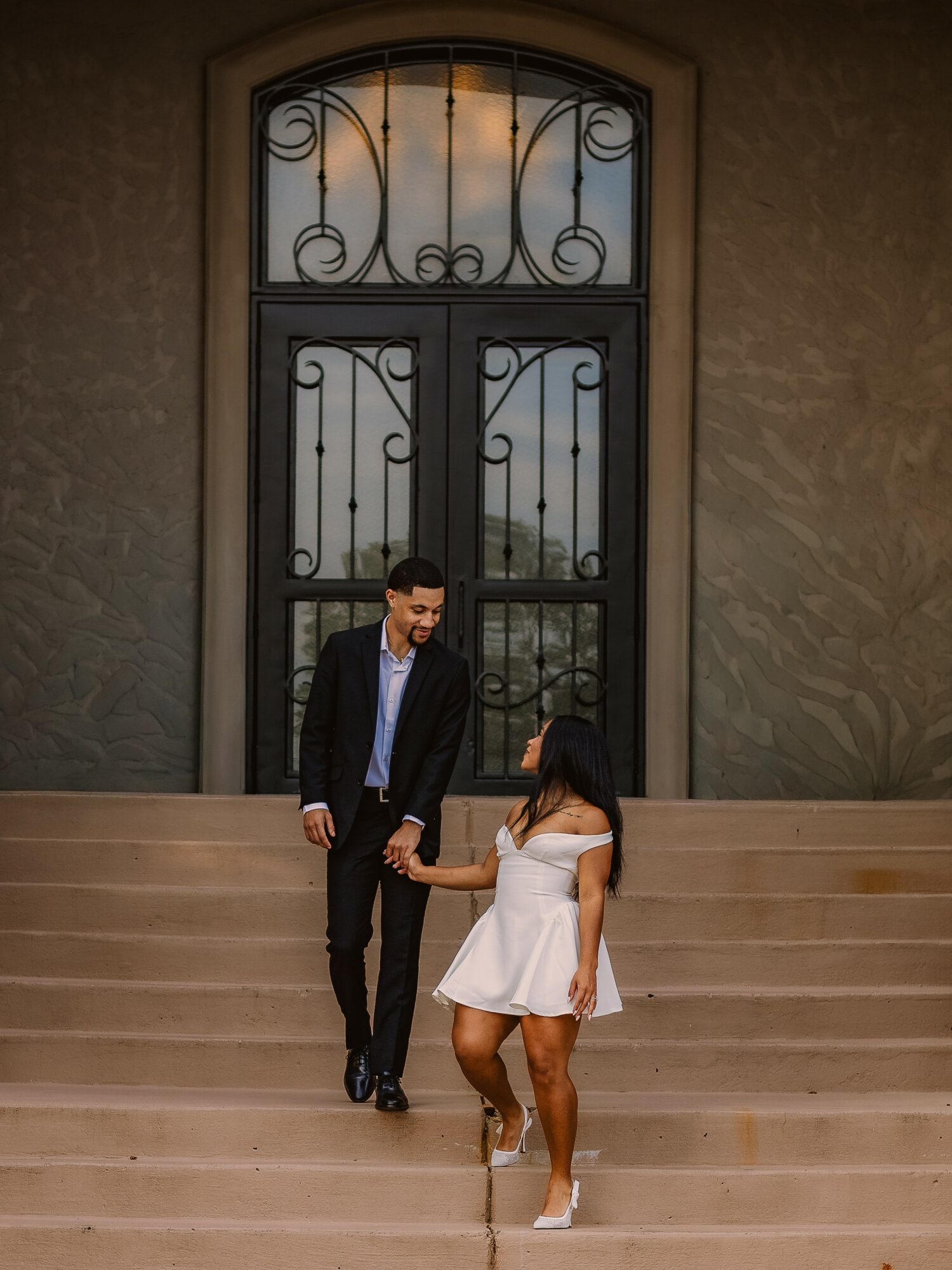 A man and woman holding hands on stairs in front of a large decorative door, woman in white dress, man in suit.