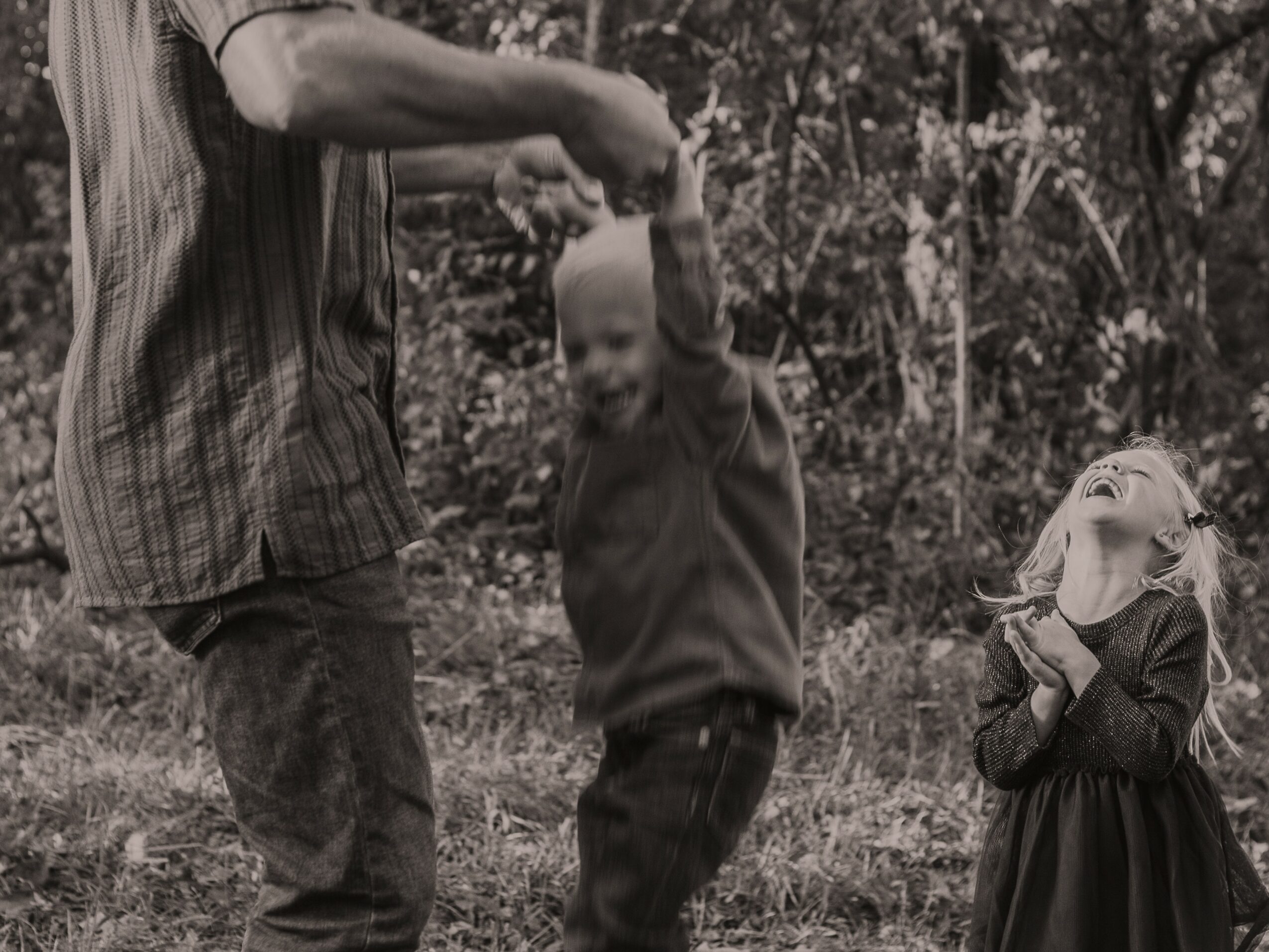 Child and adult holding hands outdoors, girl laughing and clapping, trees in background.