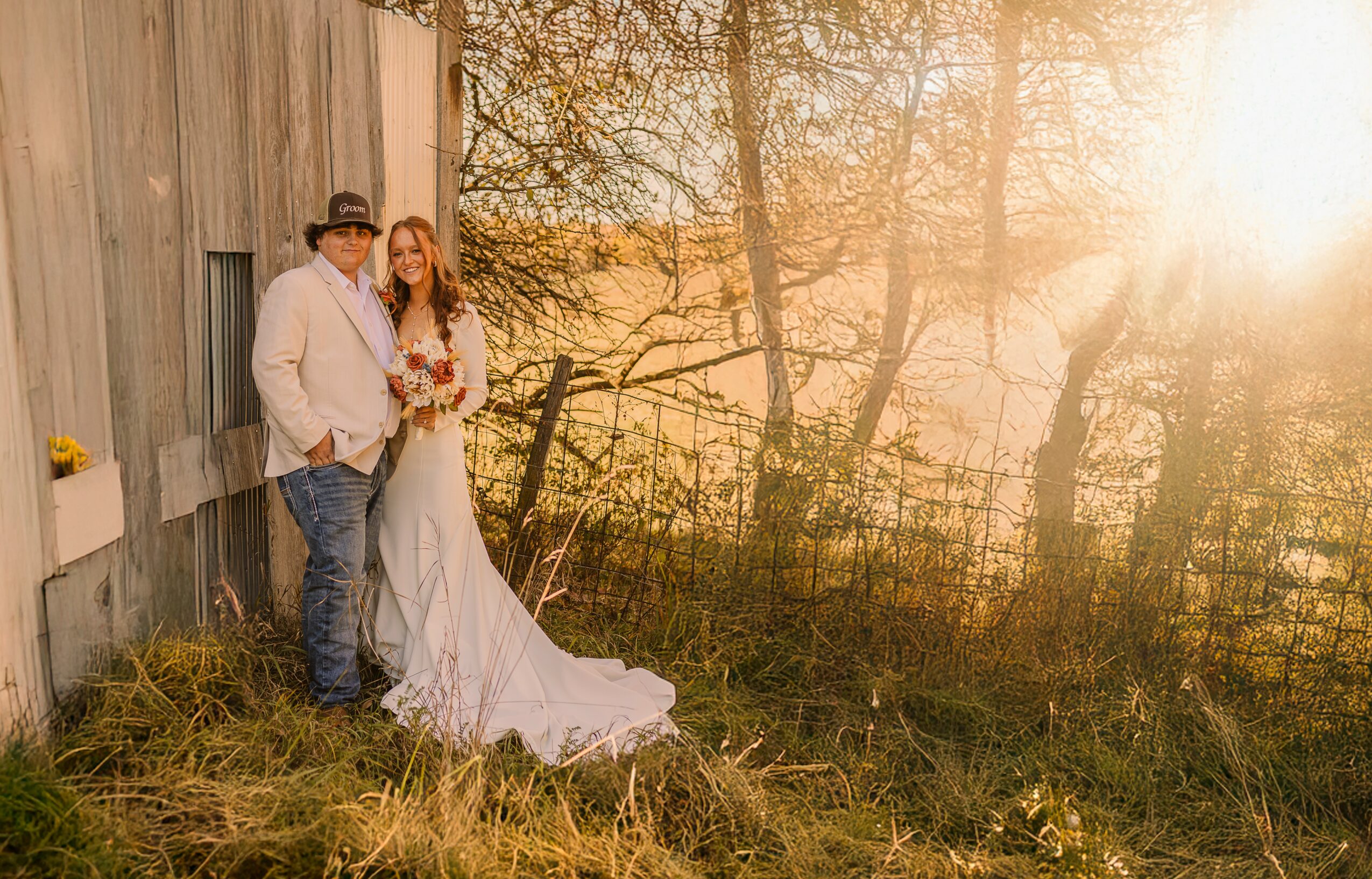 A couple in wedding attire standing outdoors near a rustic building, with sunlight filtering through trees in the background.