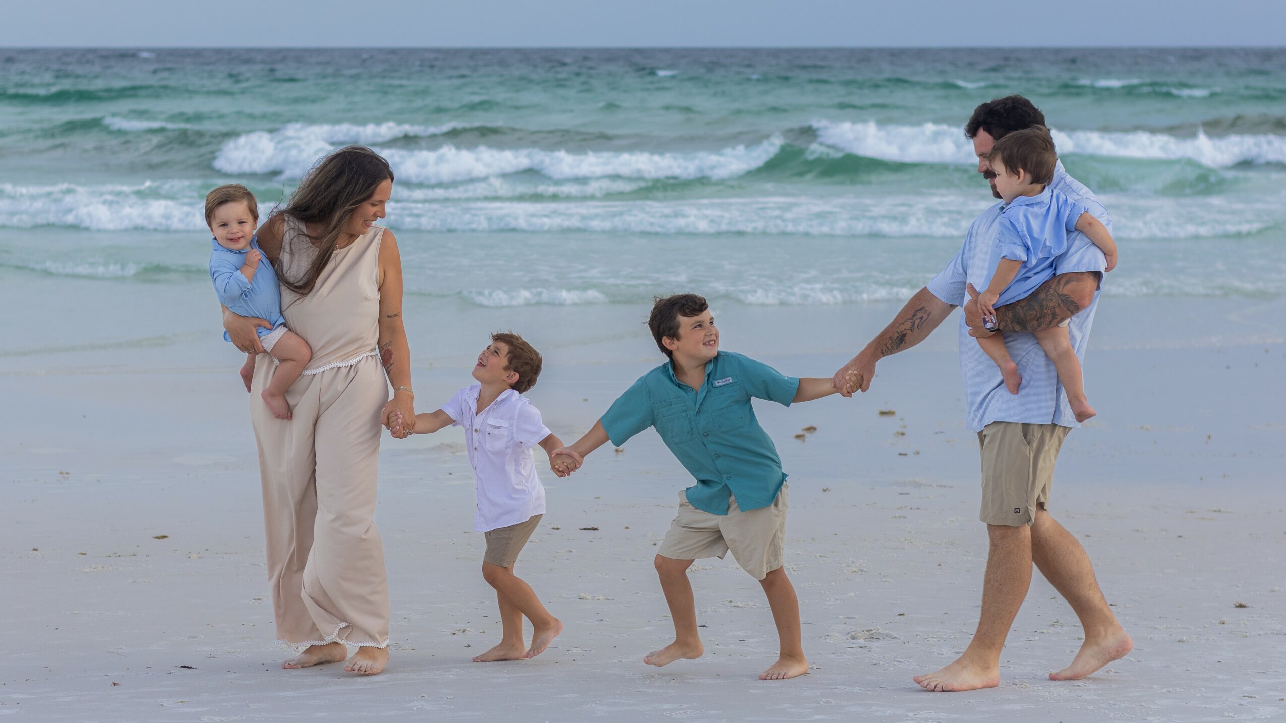 Family of six on the beach holding hands, playing near the water, with waves in the background.