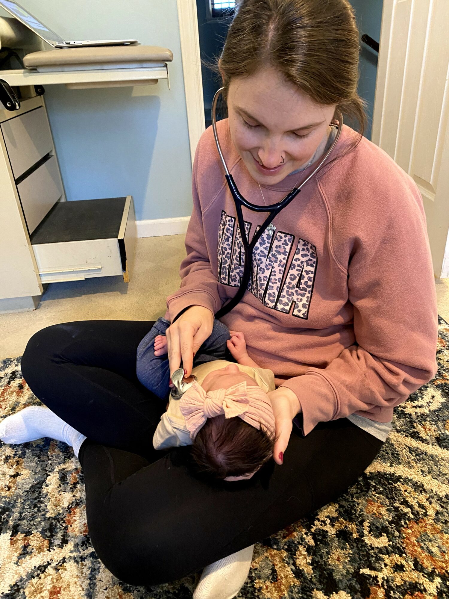 Woman sitting on a rug holding a newborn baby, wearing a stethoscope around her neck, in a room with medical equipment.