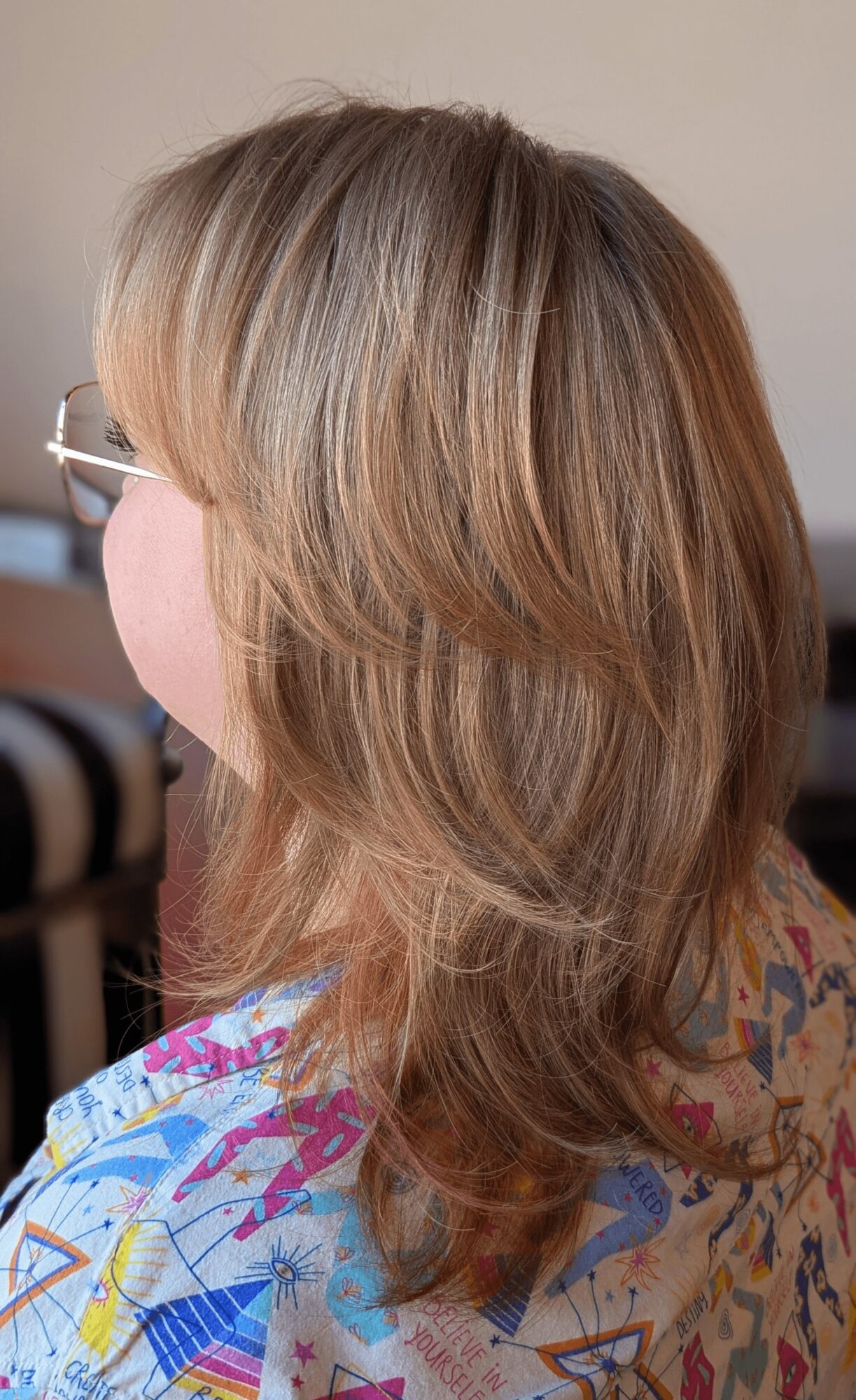 Side view of a woman with shoulder-length, wavy blonde hair wearing glasses and a colorful patterned top.