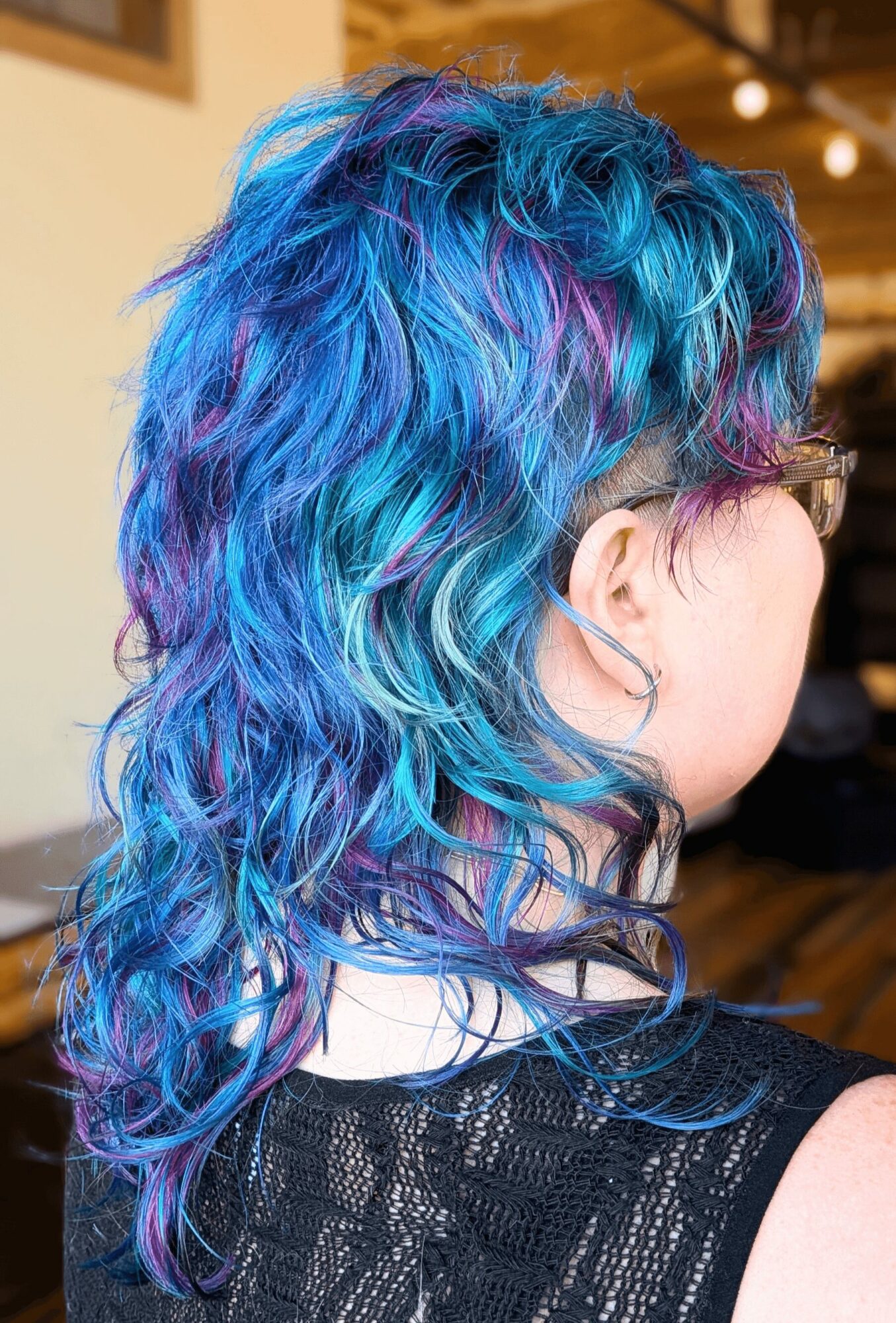 Side view of a person with vibrant, curly blue, purple, and teal hair, wearing glasses and a black top.
