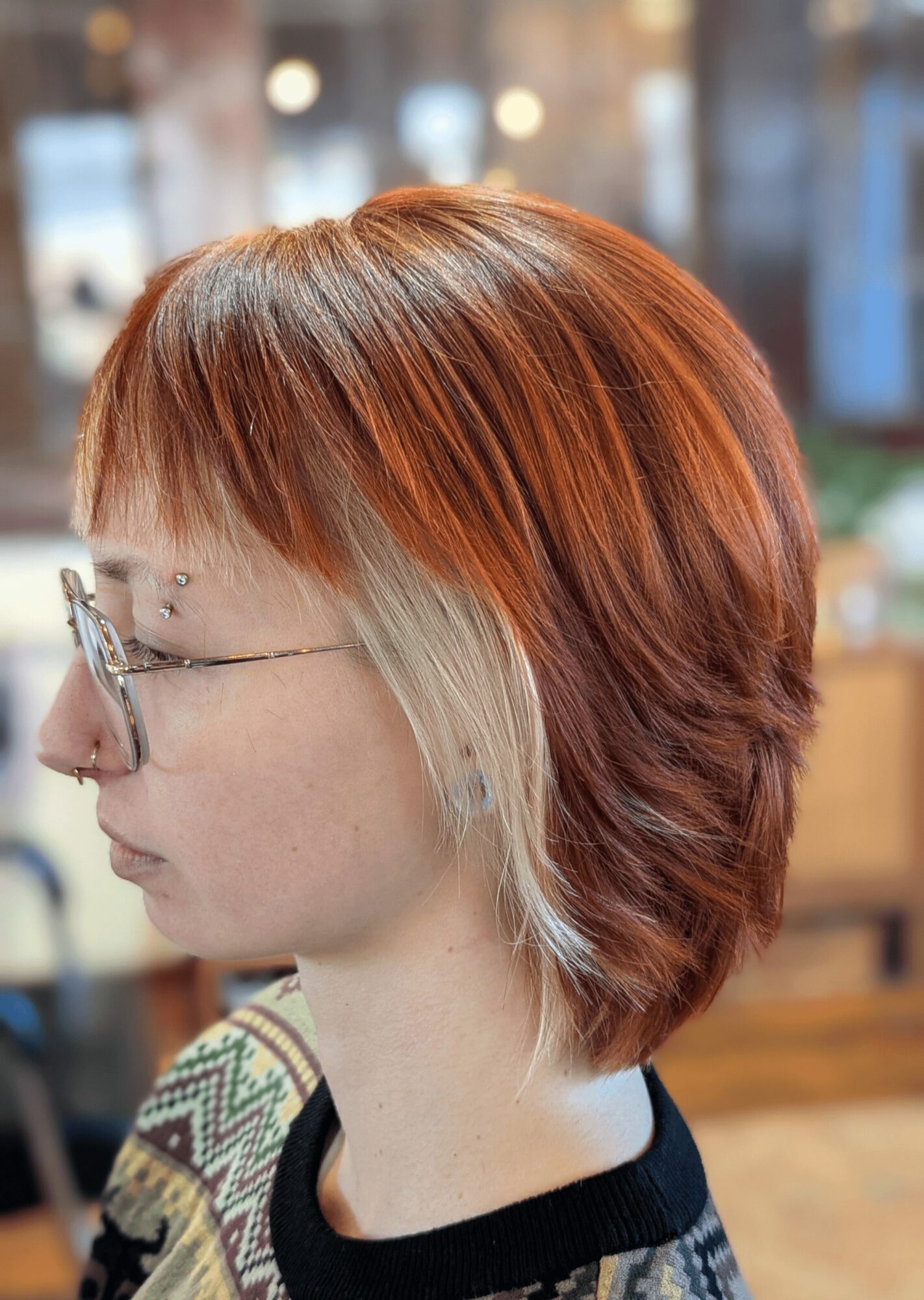 Side view of a person with short, layered hair dyed in shades of red, orange, and blonde, wearing glasses.