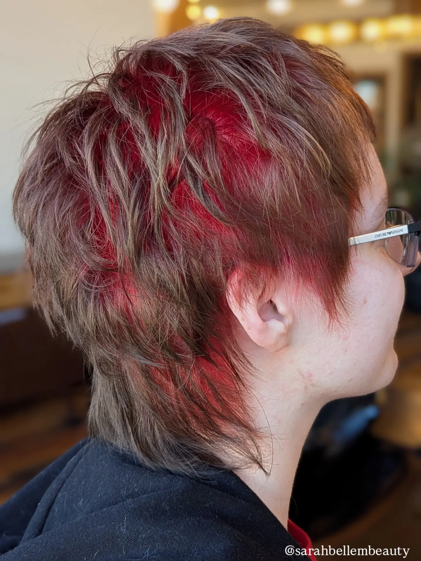 Side view of a person with short, layered hair dyed red and brown, wearing glasses, indoors.