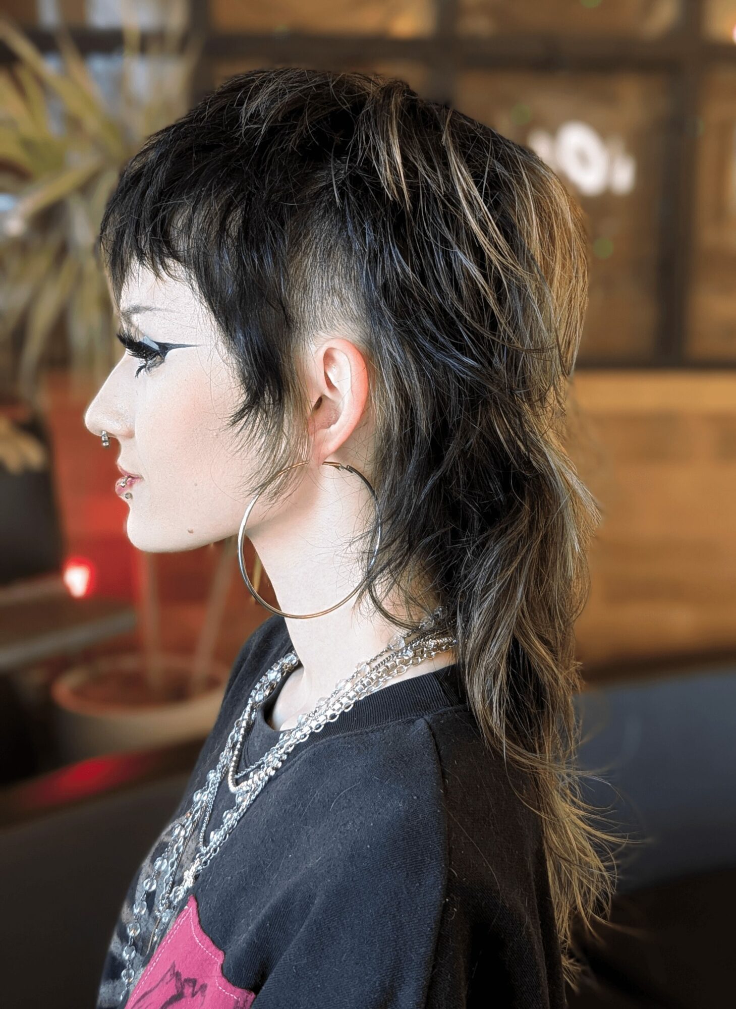 Side profile of a woman with short, layered black hair and a mullet style, wearing jewelry, in an indoor setting.