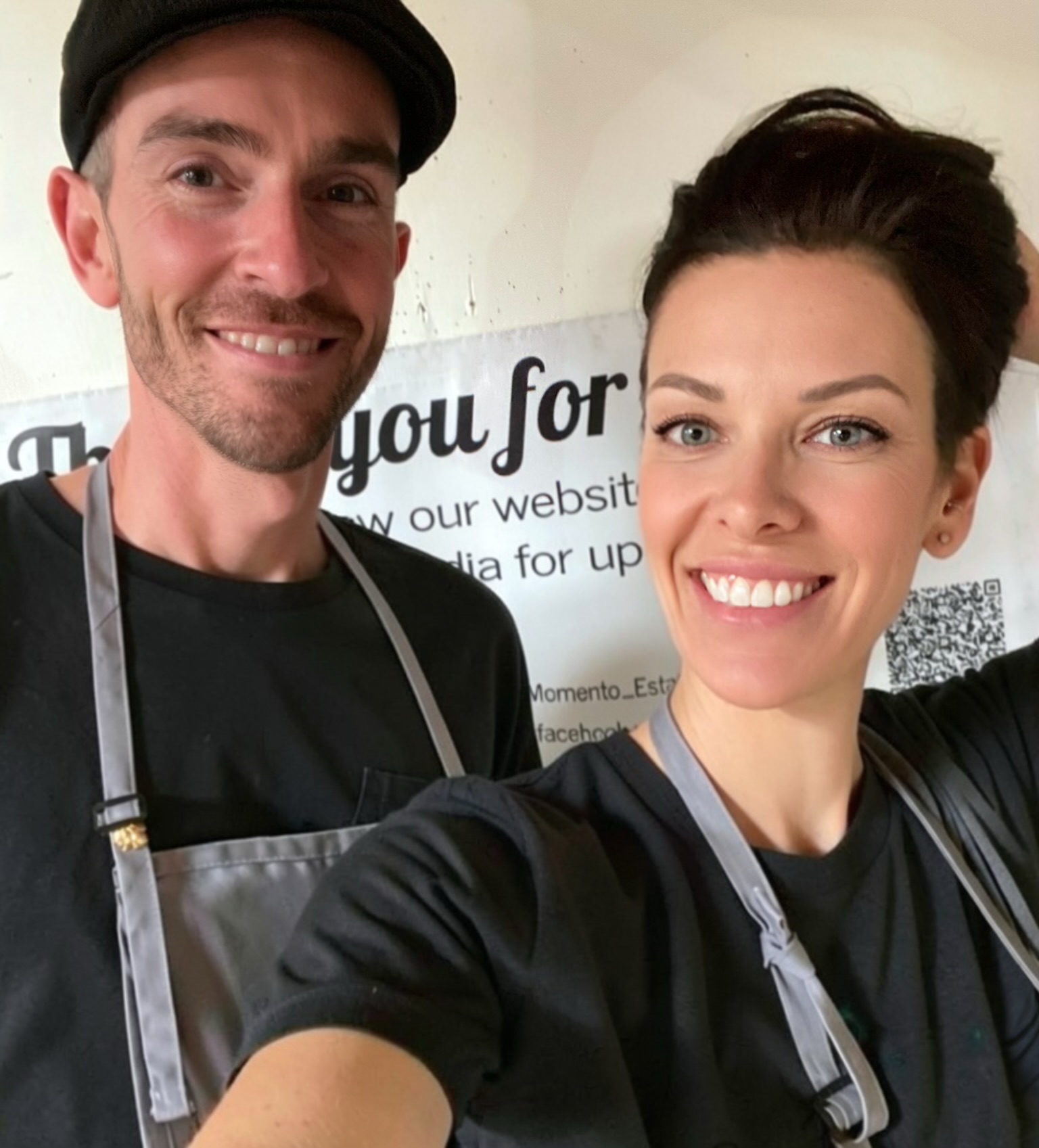 Two smiling people taking a selfie, standing in front of a sign with text, both wearing black shirts and aprons.