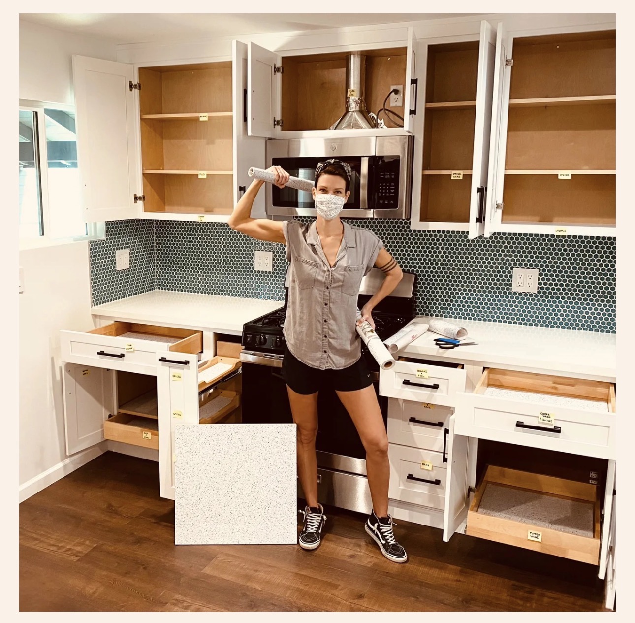 Person wearing a mask, holding a tool, standing in a kitchen with open cabinets and drawers, wooden floor, and blue backsplash.