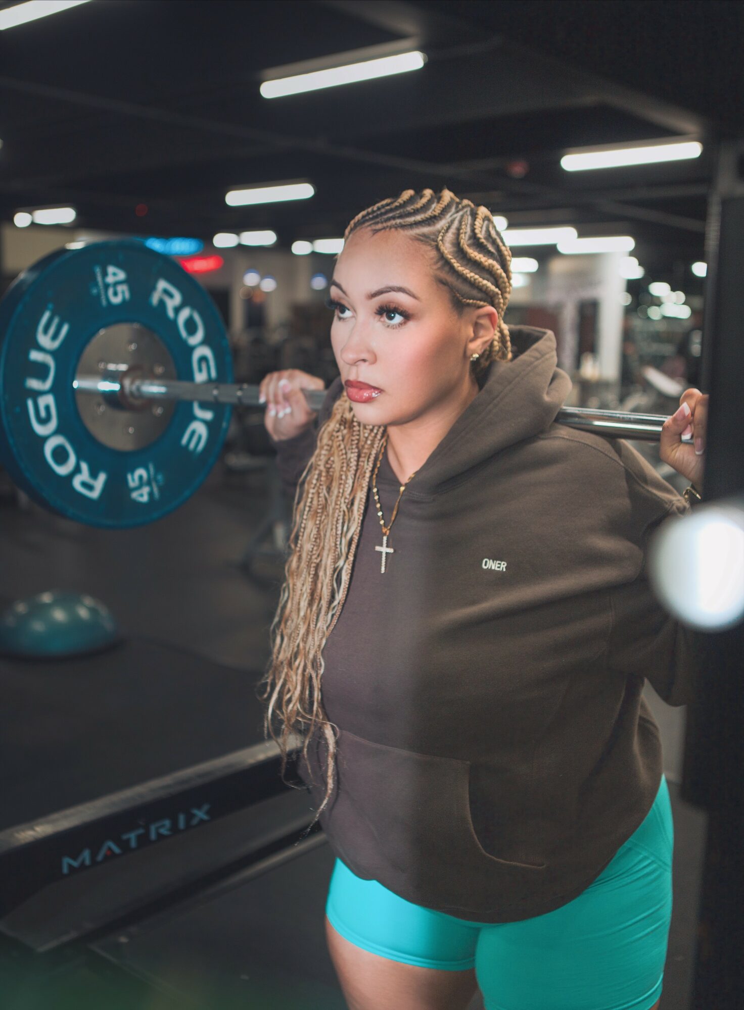 Woman with braided hair lifting weights in a gym, wearing a black hoodie and turquoise shorts, focused expression.
